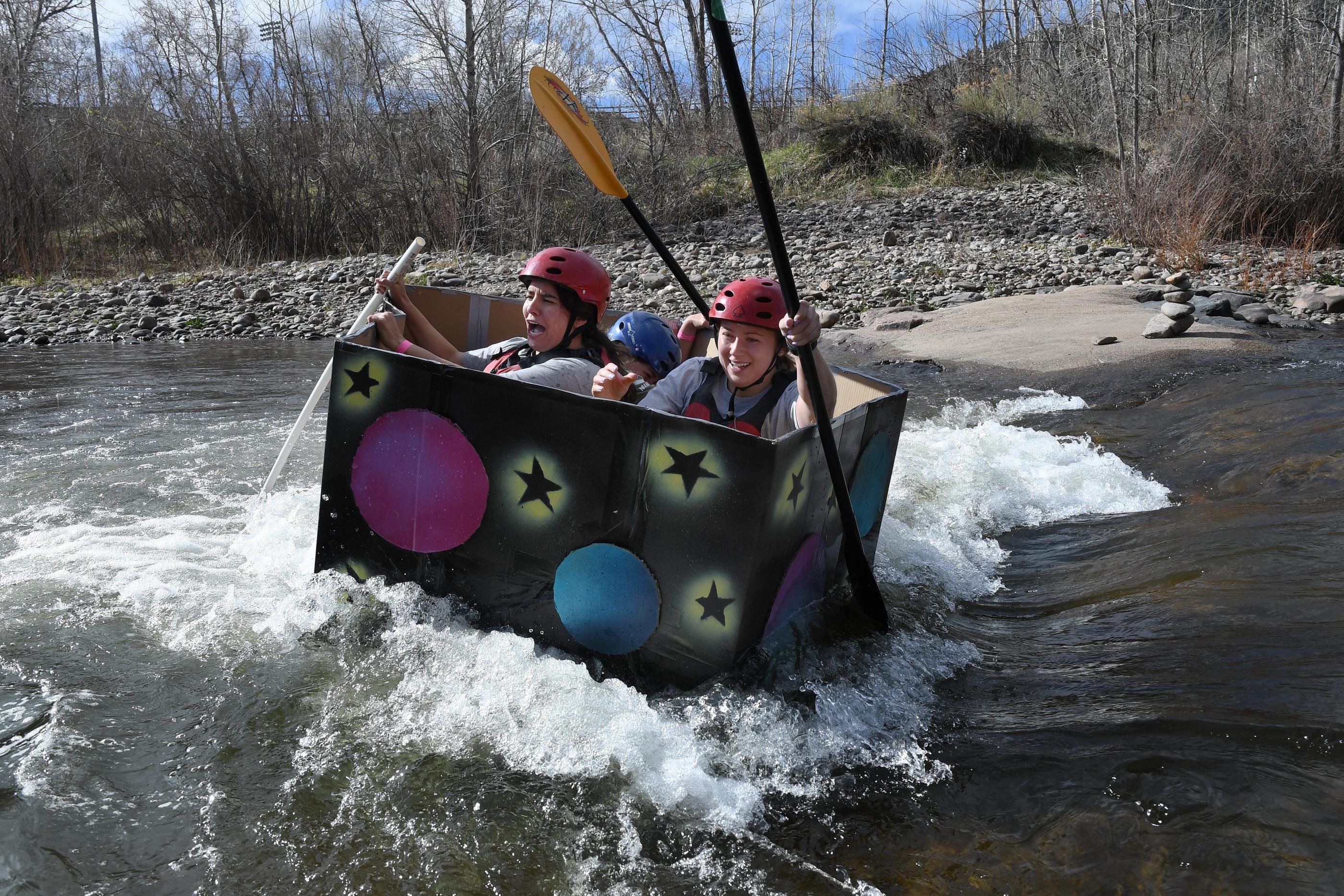 Students in red helmets row a cardboard boat