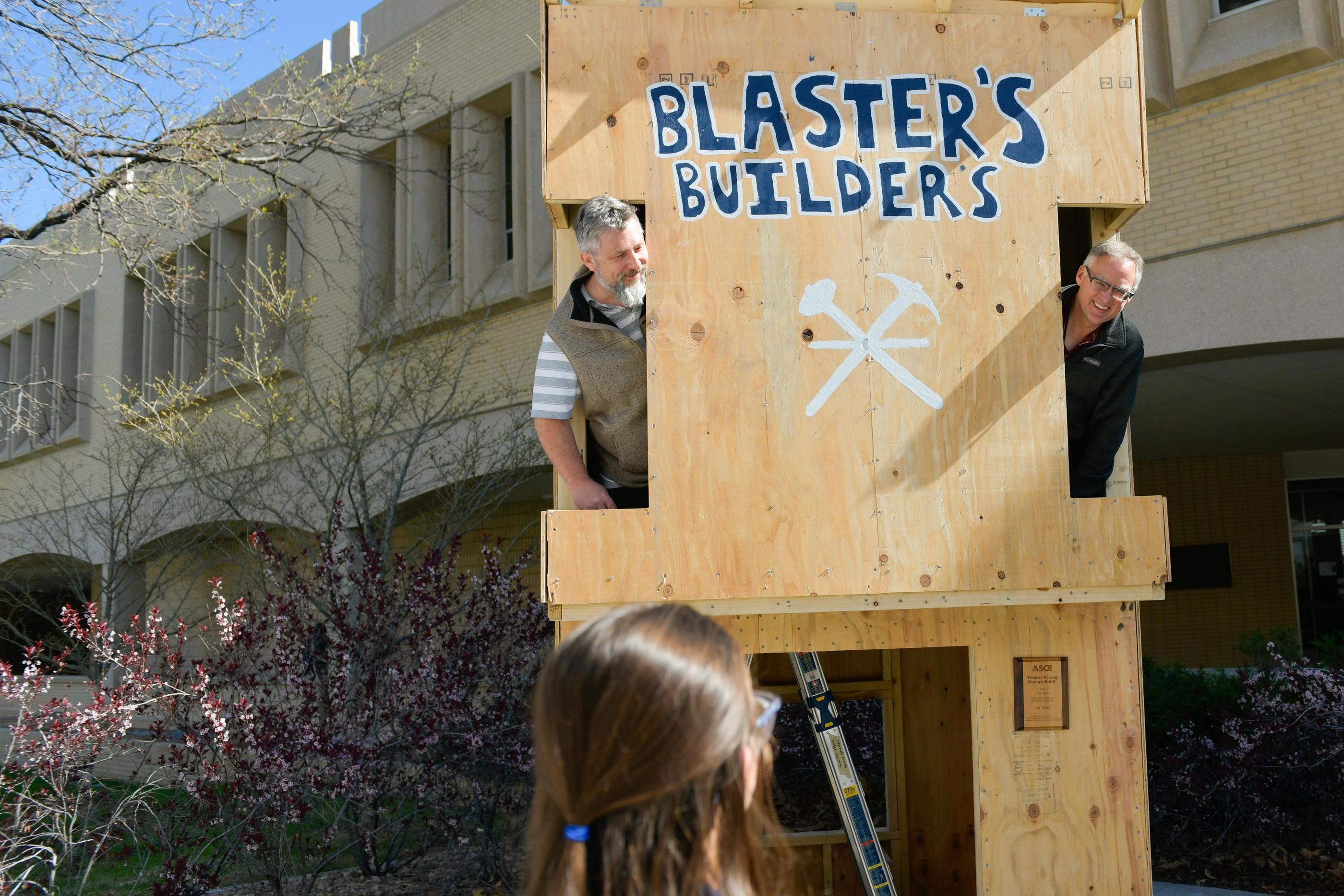 The heads of two men poke out the windows of a two-story timber-built building painted with the name Blaster's Building