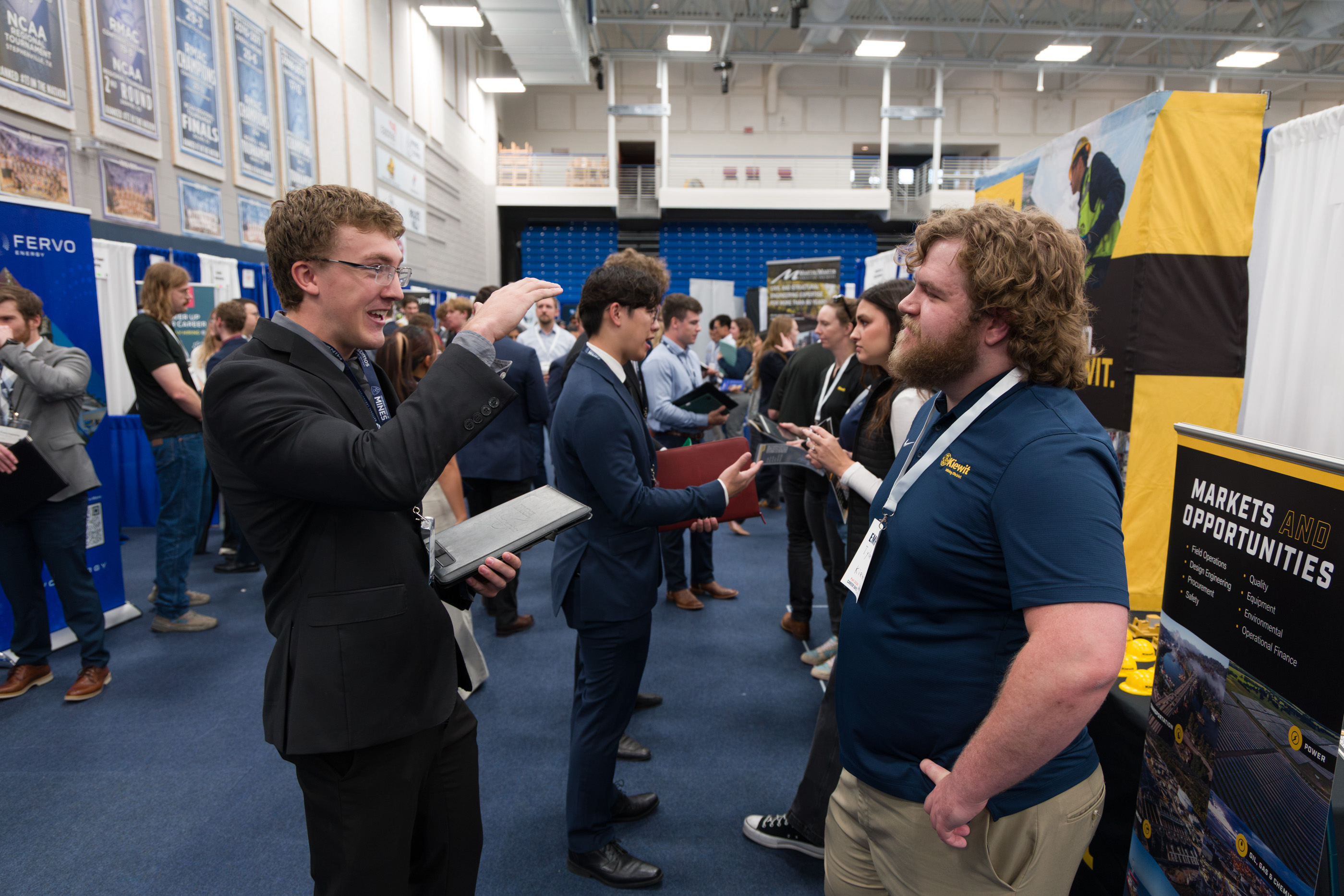 A student in a black suit gestures while talking with a bearded Kiewit recruiter beside a yellow and black booth