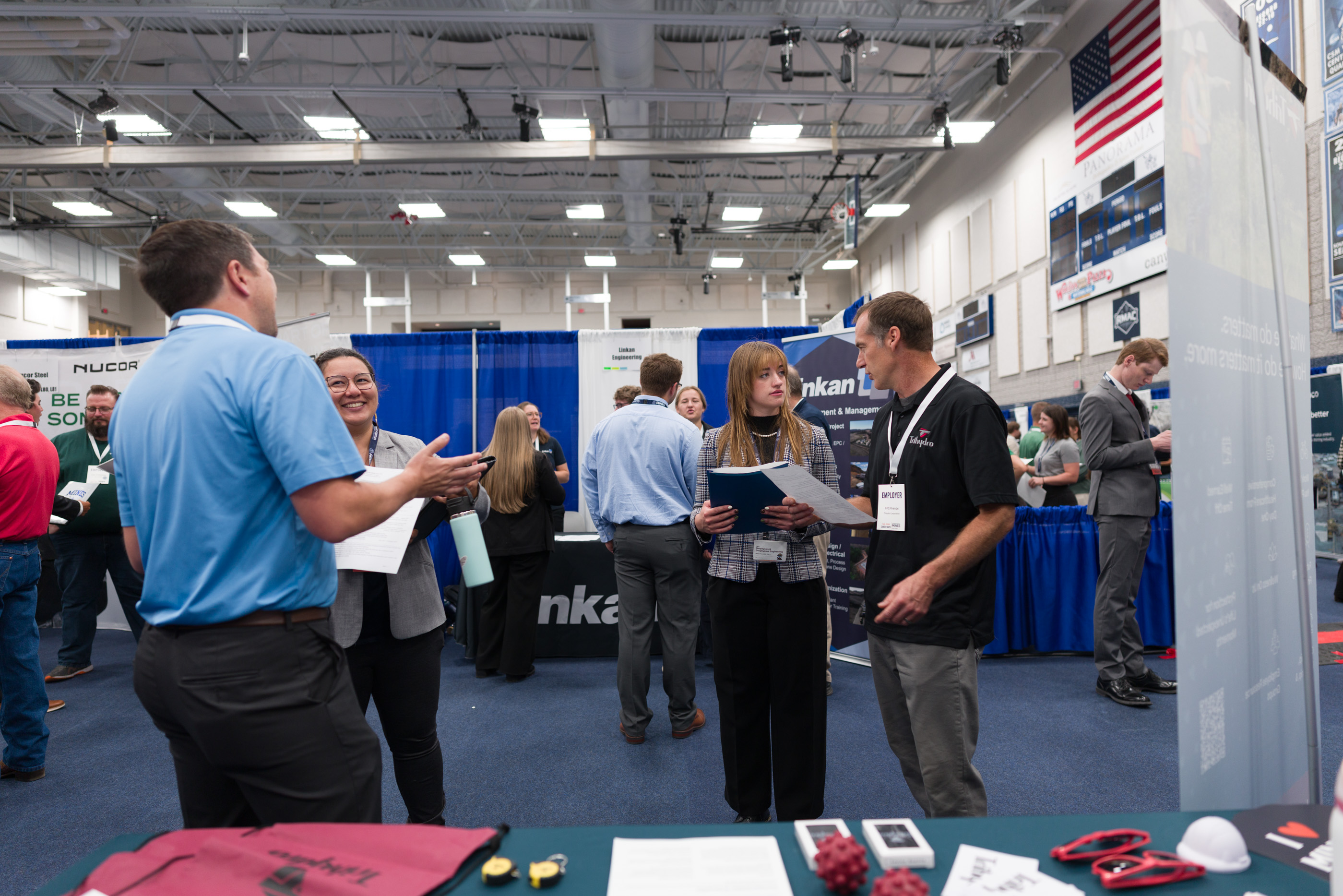 A student in a plaid blazer reviews papers with a Trihydro recruiter as another student smiles while talking with a recruiter nearby in a gym filled with booths