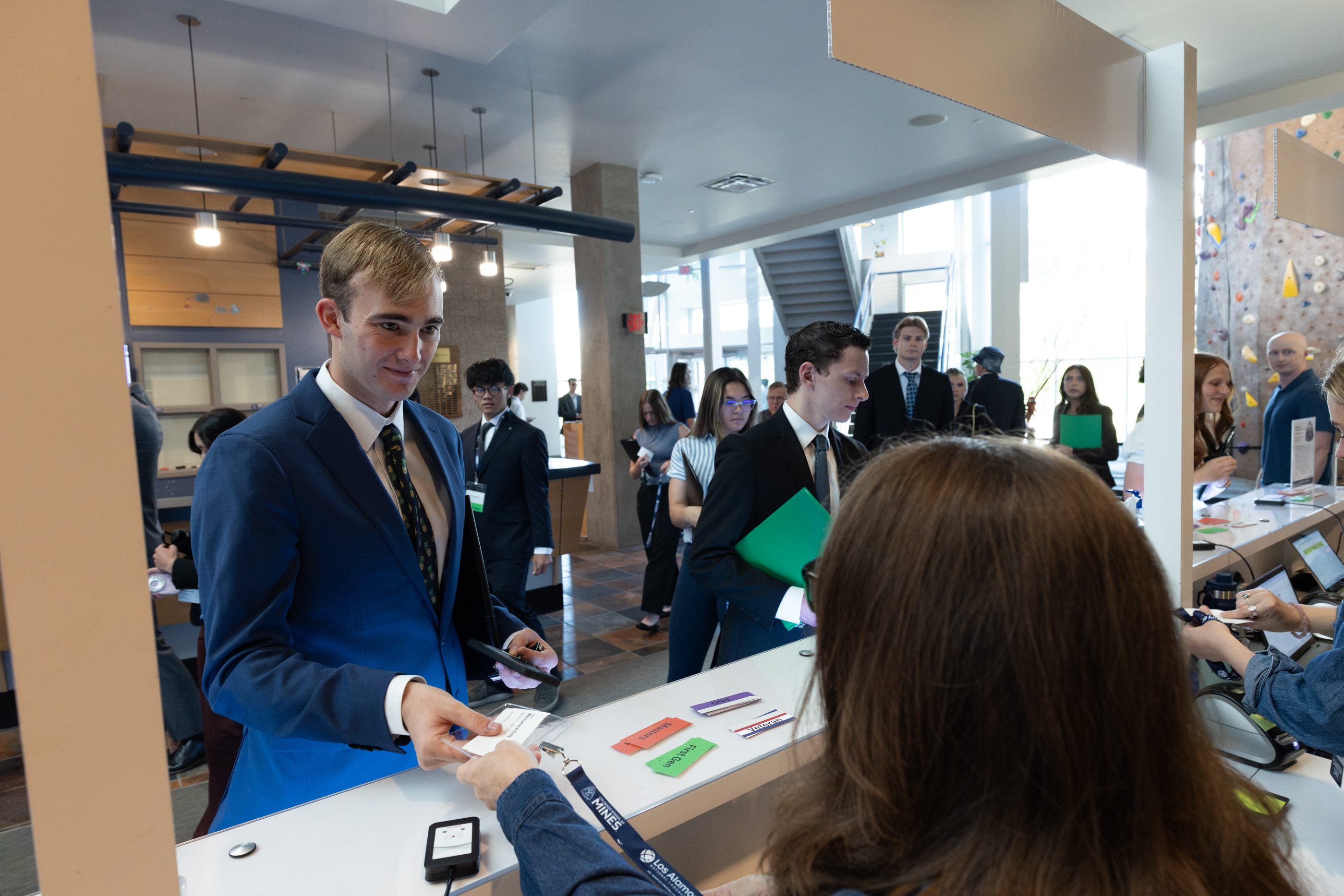 A student in a blue suit hands his ID to a staff member at a check-in counter as students in business attire wait in line behind him