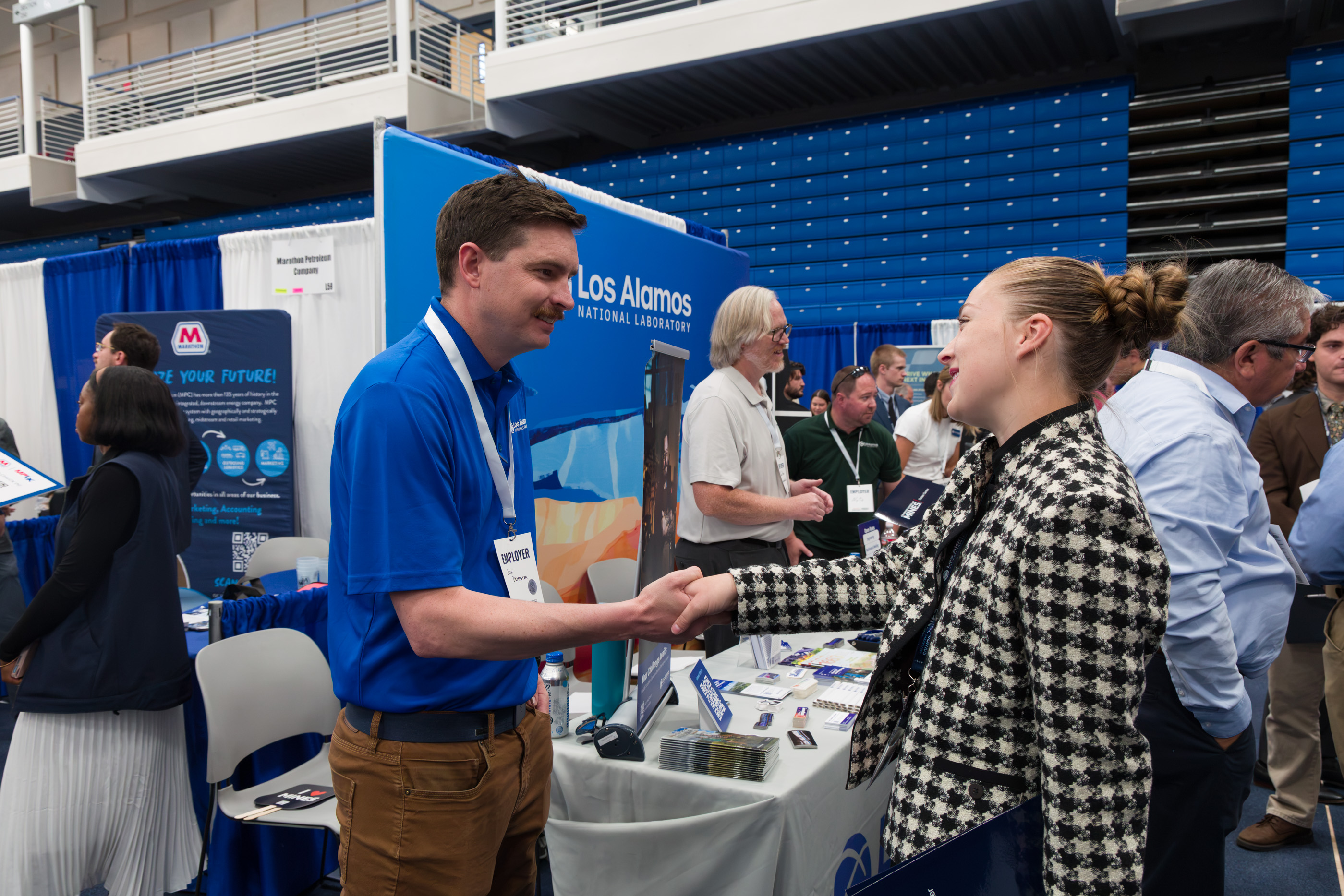 A student in a houndstooth jacket shakes hands with a recruiter in a blue polo at the Los Alamos National Laboratory booth