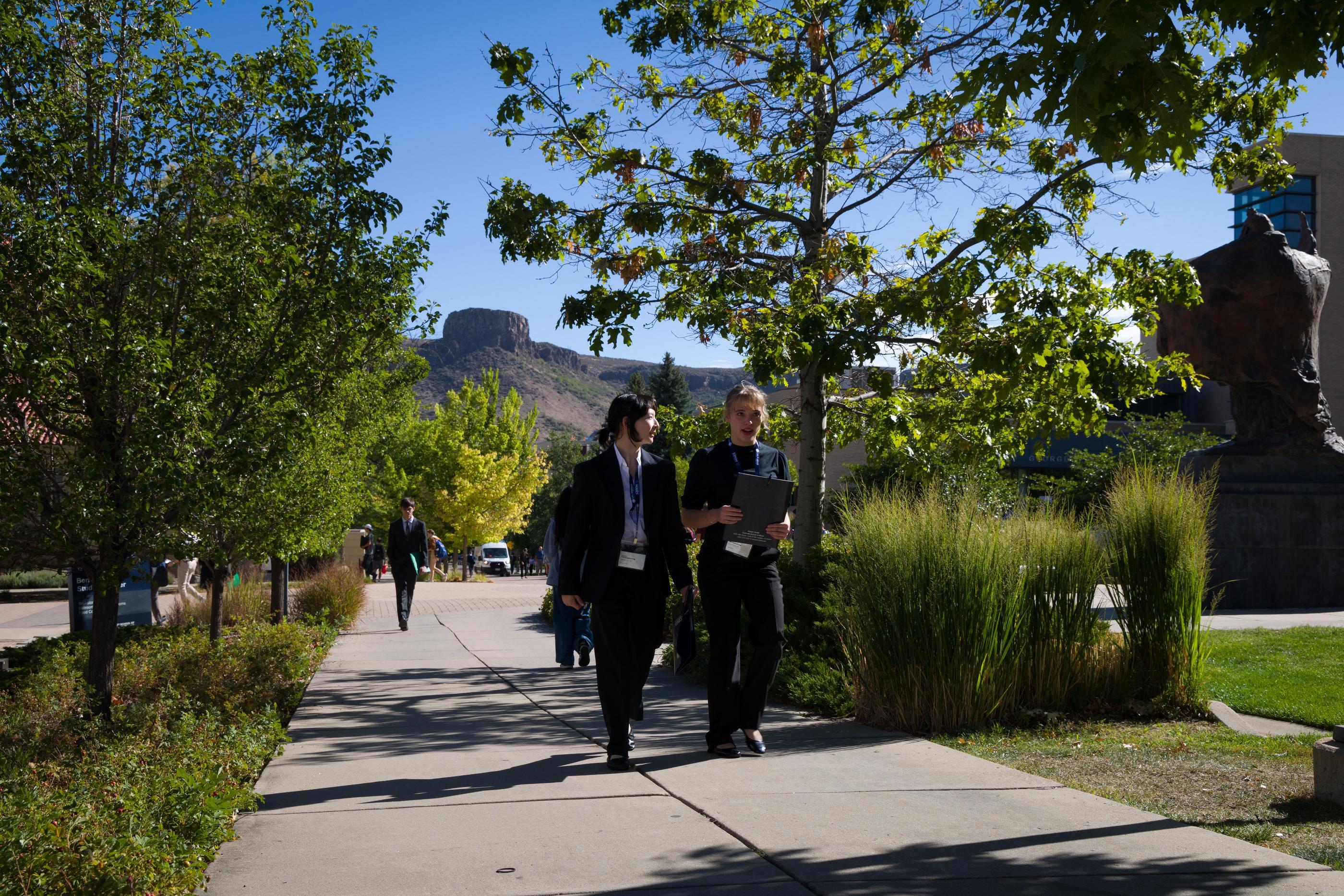 Two students in business wear walk across campus with South Table Mountain in the background
