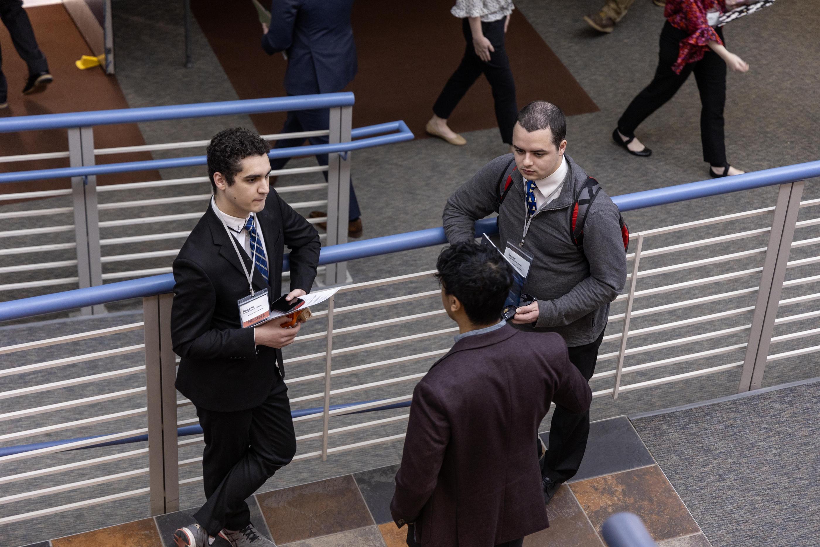 Two male students in suits lean against a railing
