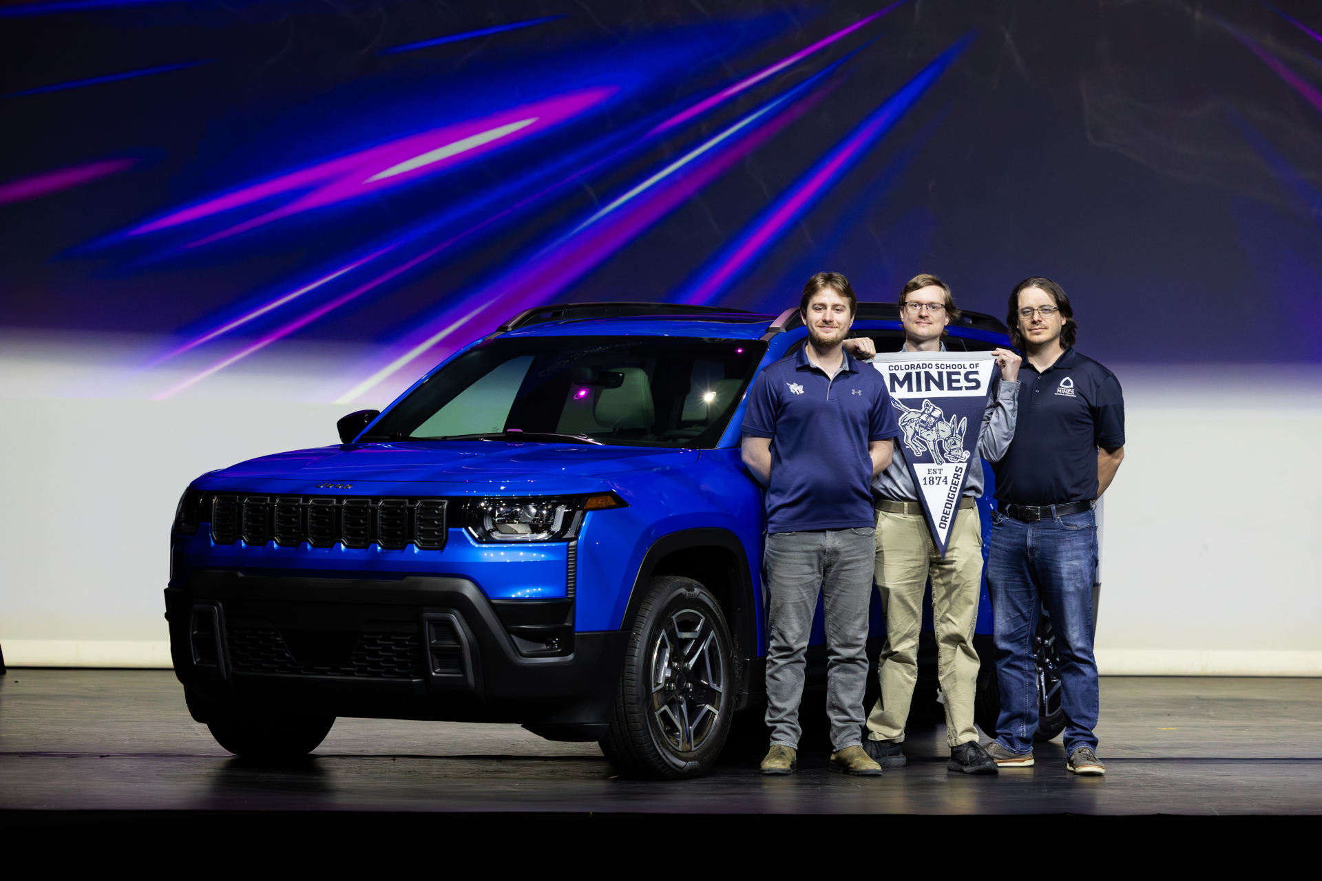 three men stand in front of a blue car holding a Mines pennant