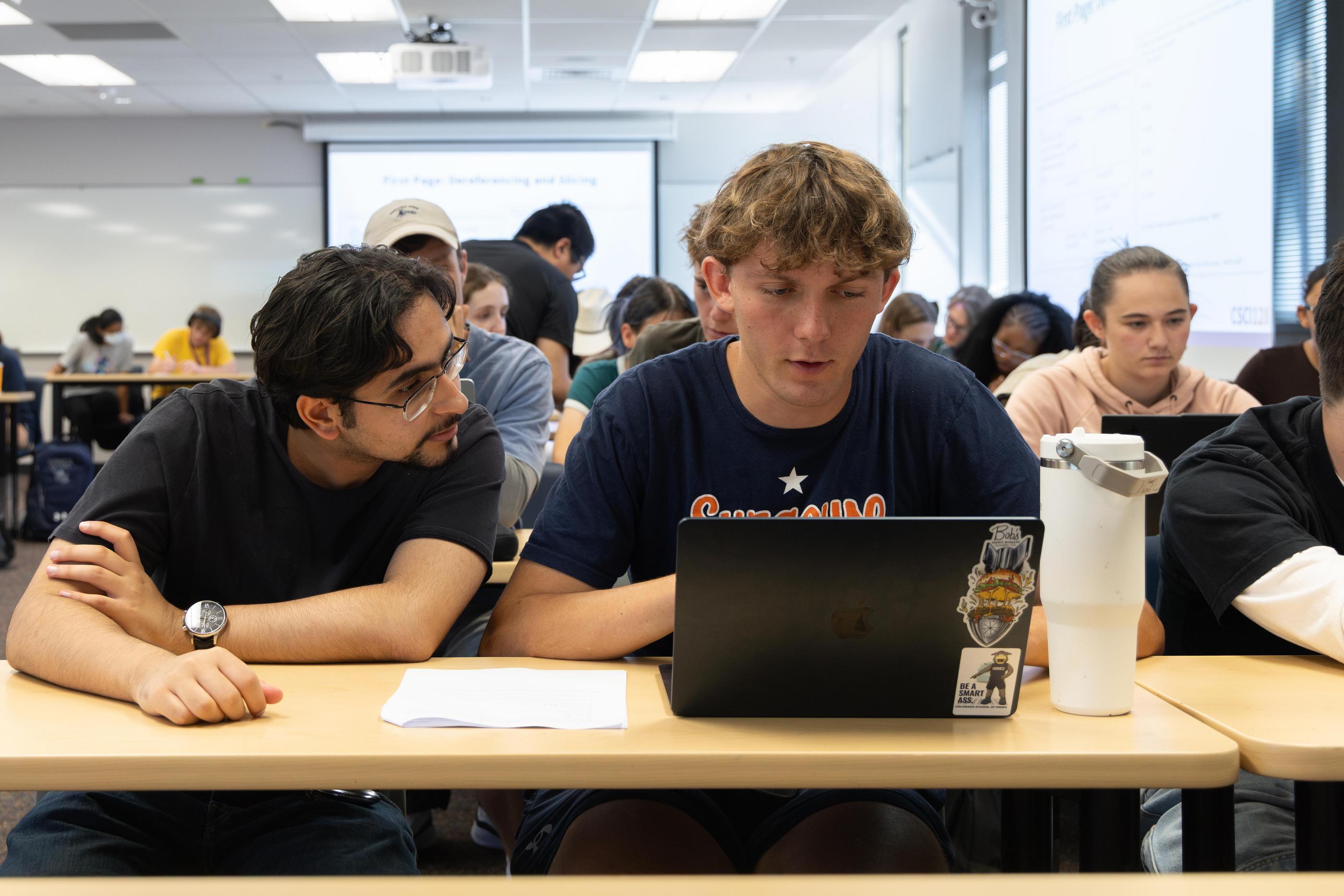 Two students look at a laptop screen on the desk in front of them during class