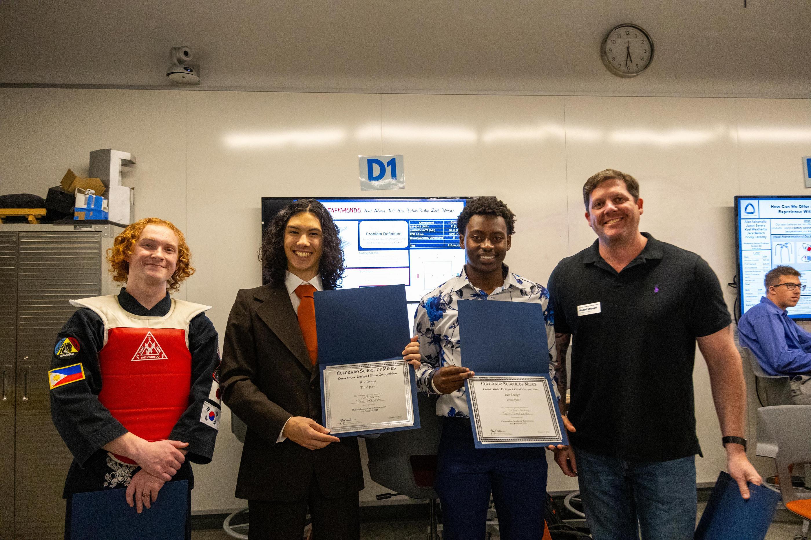 Two students hold award certificates next to student wearing prototype protective vest