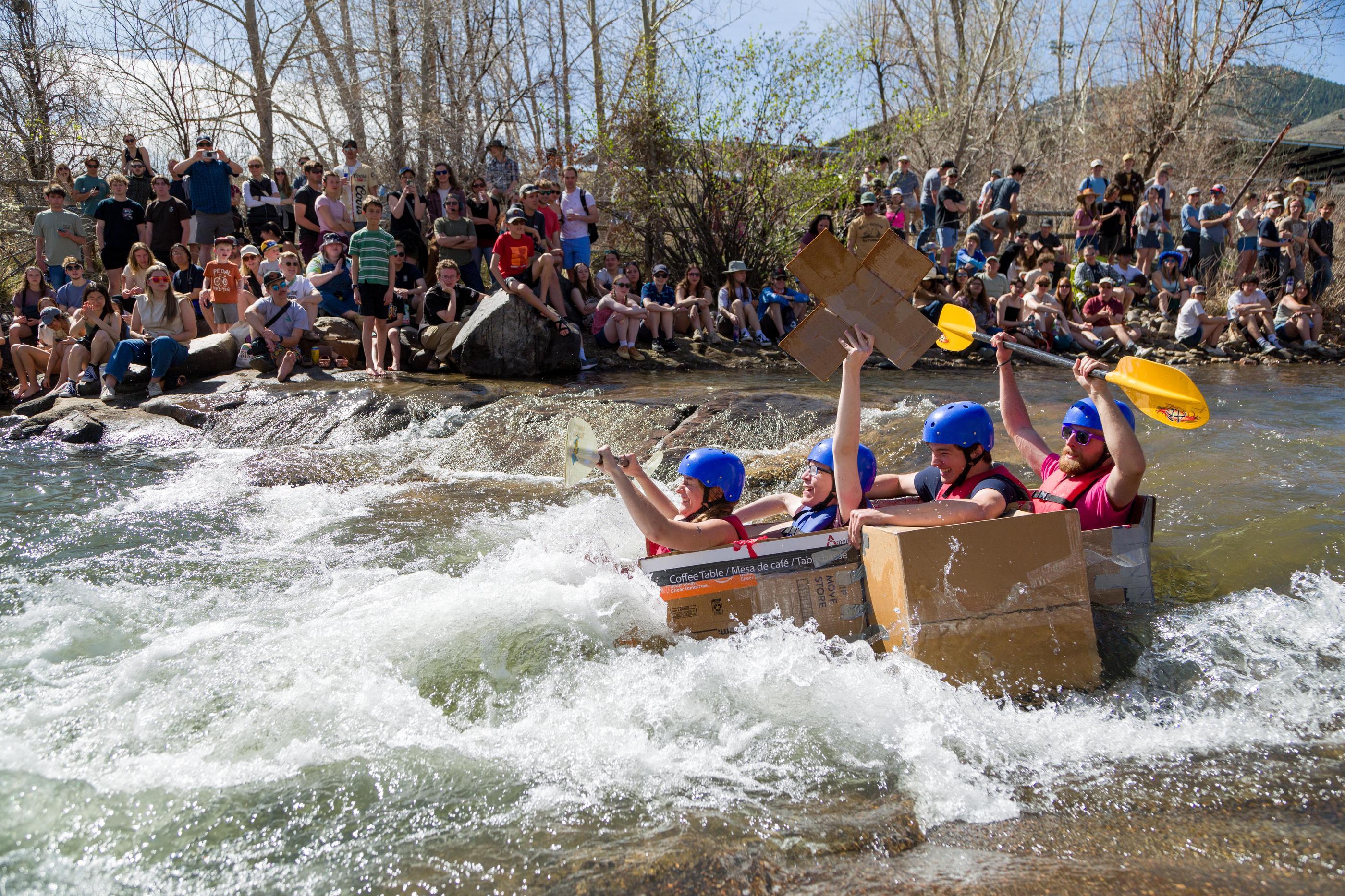 Four students paddle a cardboard boat through whitewater rapids on Clear Creek during the Colorado School of Mines E-Days Cardboard Boat Race, as a large crowd watches from the bank.