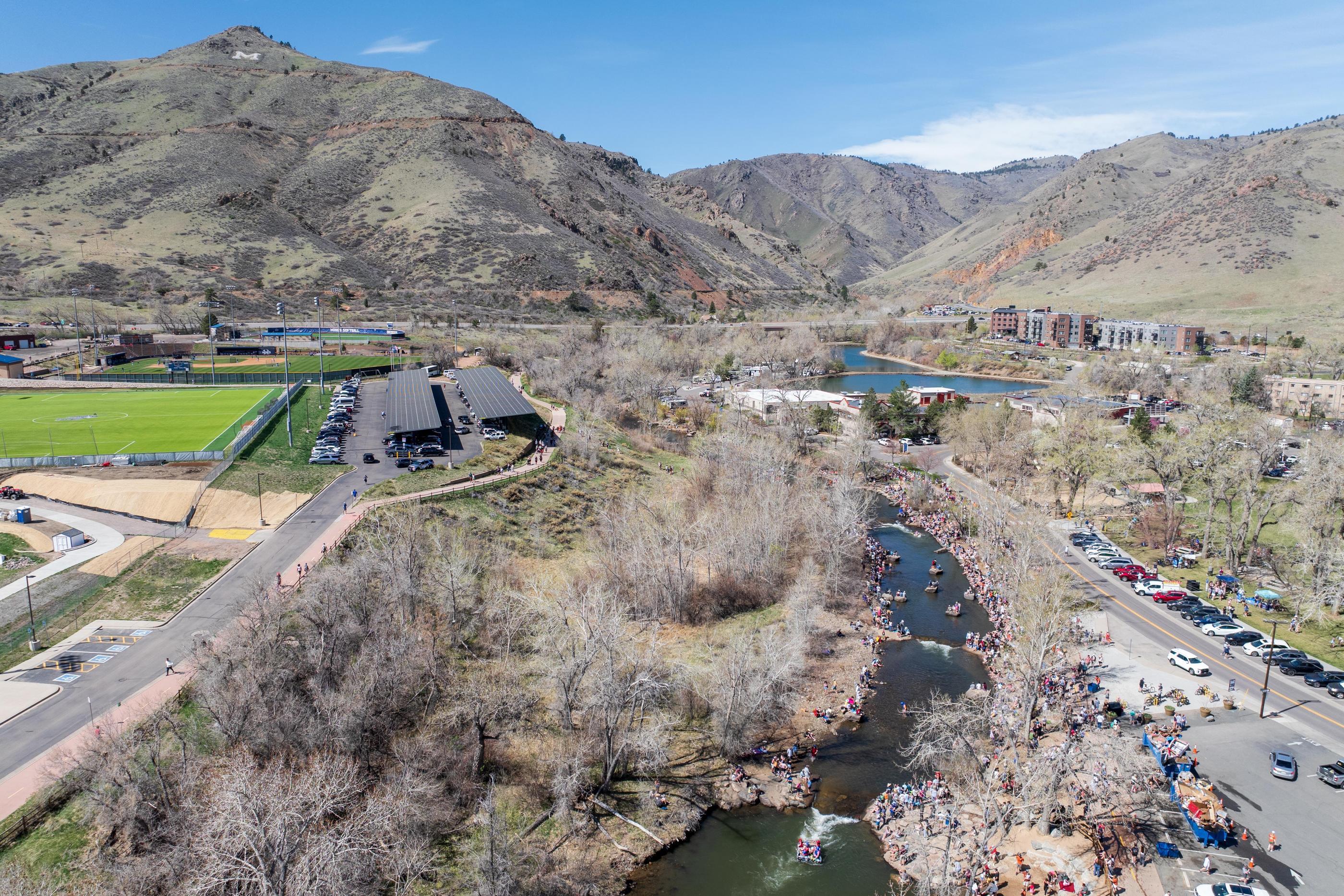 An aerial view of Clear Creek in Golden, Colorado, shows crowds lining the banks for the Colorado School of Mines Cardboard Boat Race, with campus athletic fields and the M on the mountainside in the background.