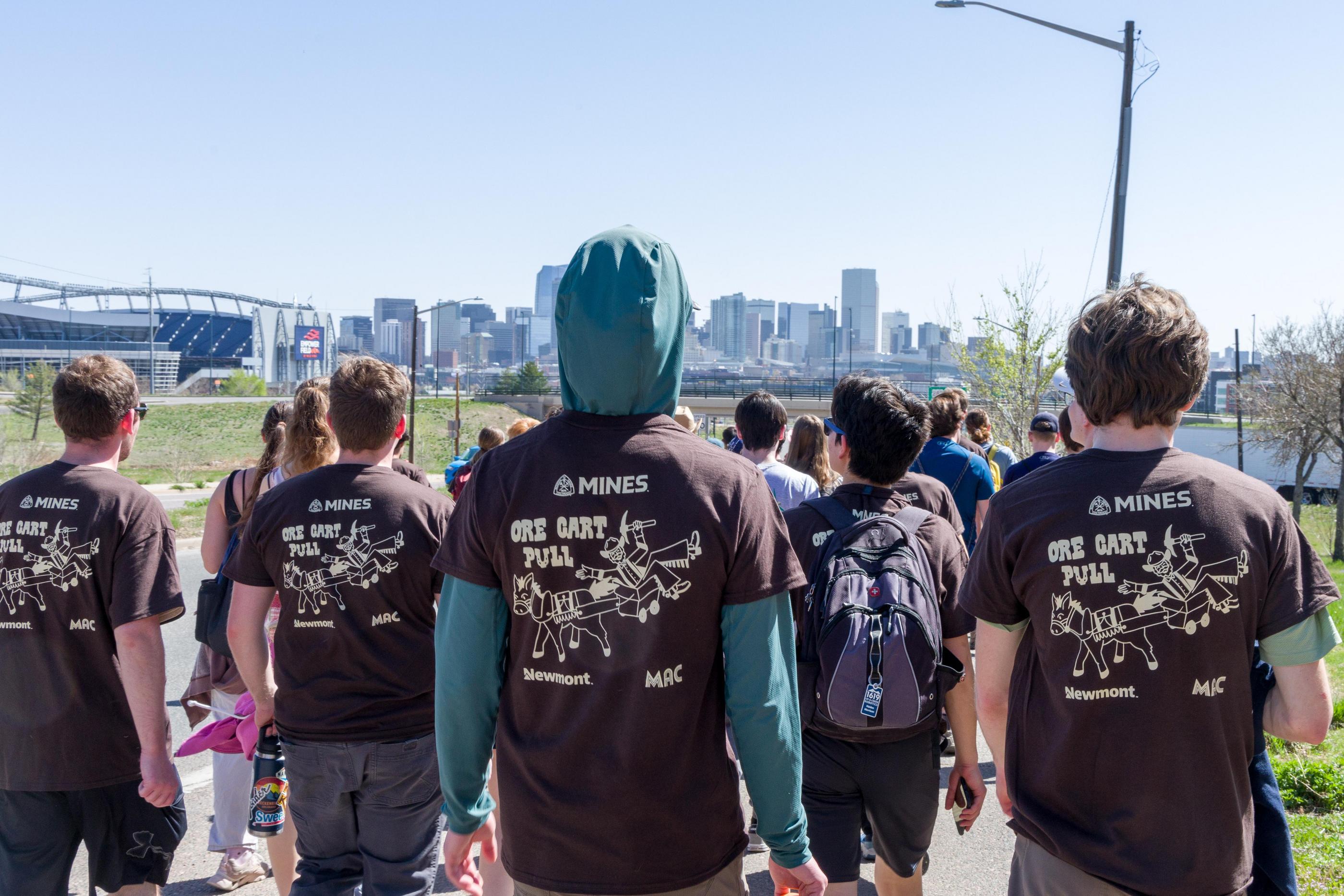 Students wearing Mines Ore Cart Pull T-shirts walk together with the Denver skyline and a football stadium in the background during E-Days.