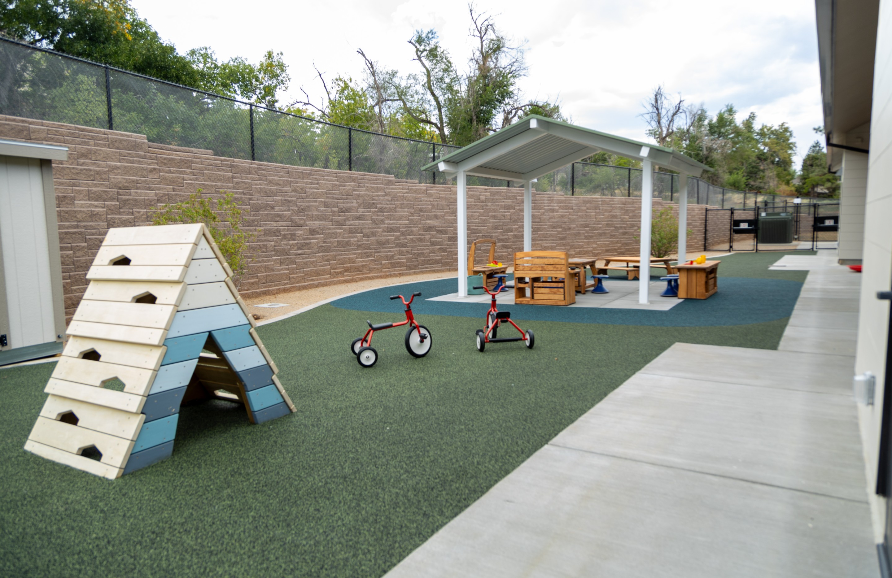 The outdoor play yard at the Mines Early Childhood Center with a wooden climbing structure, tricycles on turf, and a covered picnic area against a stone wall.
