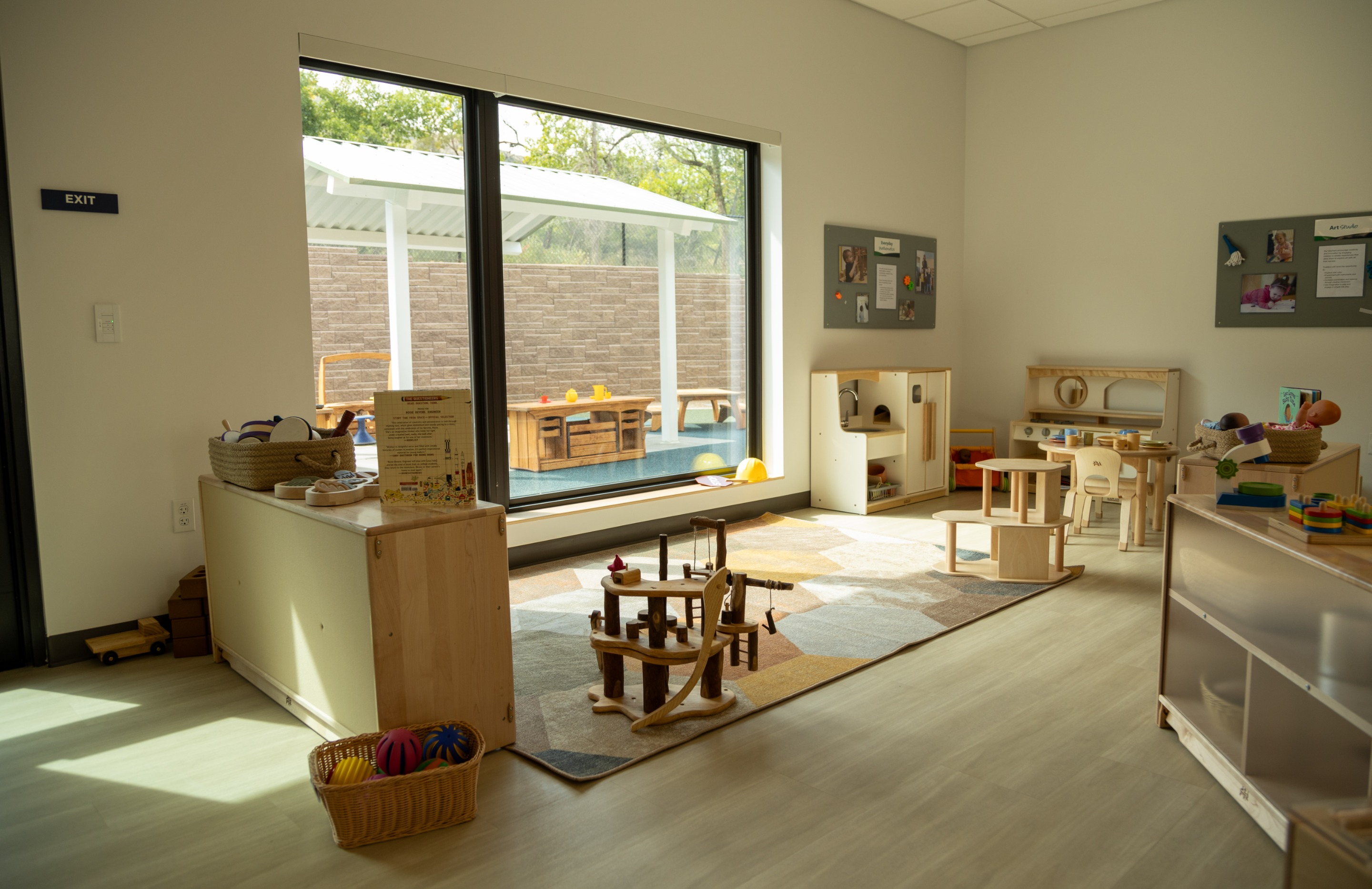 A classroom nook at the Mines Early Childhood Center with a play kitchen, wooden rocking toy, low tables and chairs, and a large window looking out to a covered outdoor patio.