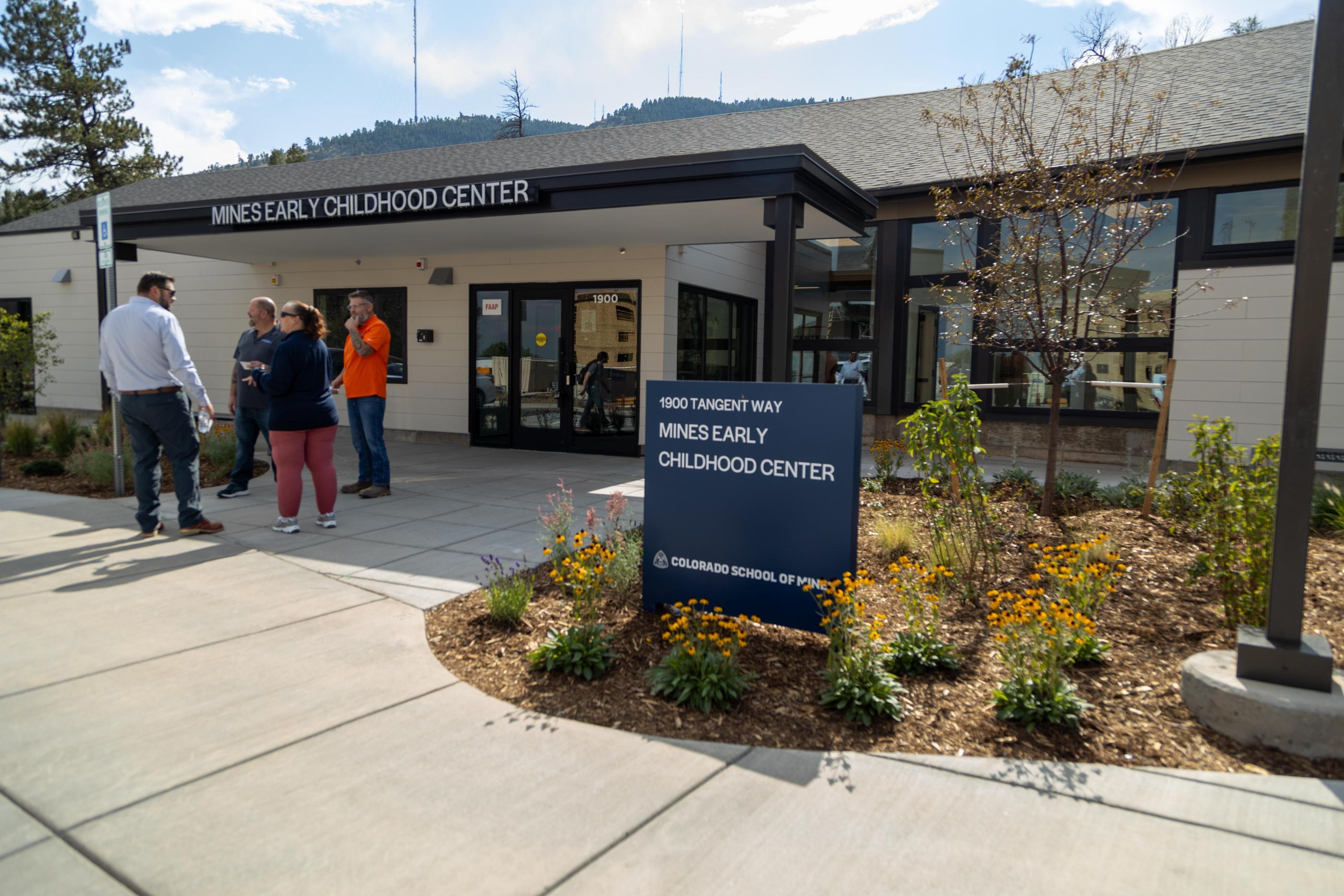 Four adults talk outside the entrance of the Mines Early Childhood Center, with the building's sign and landscaped entry visible in the foreground.