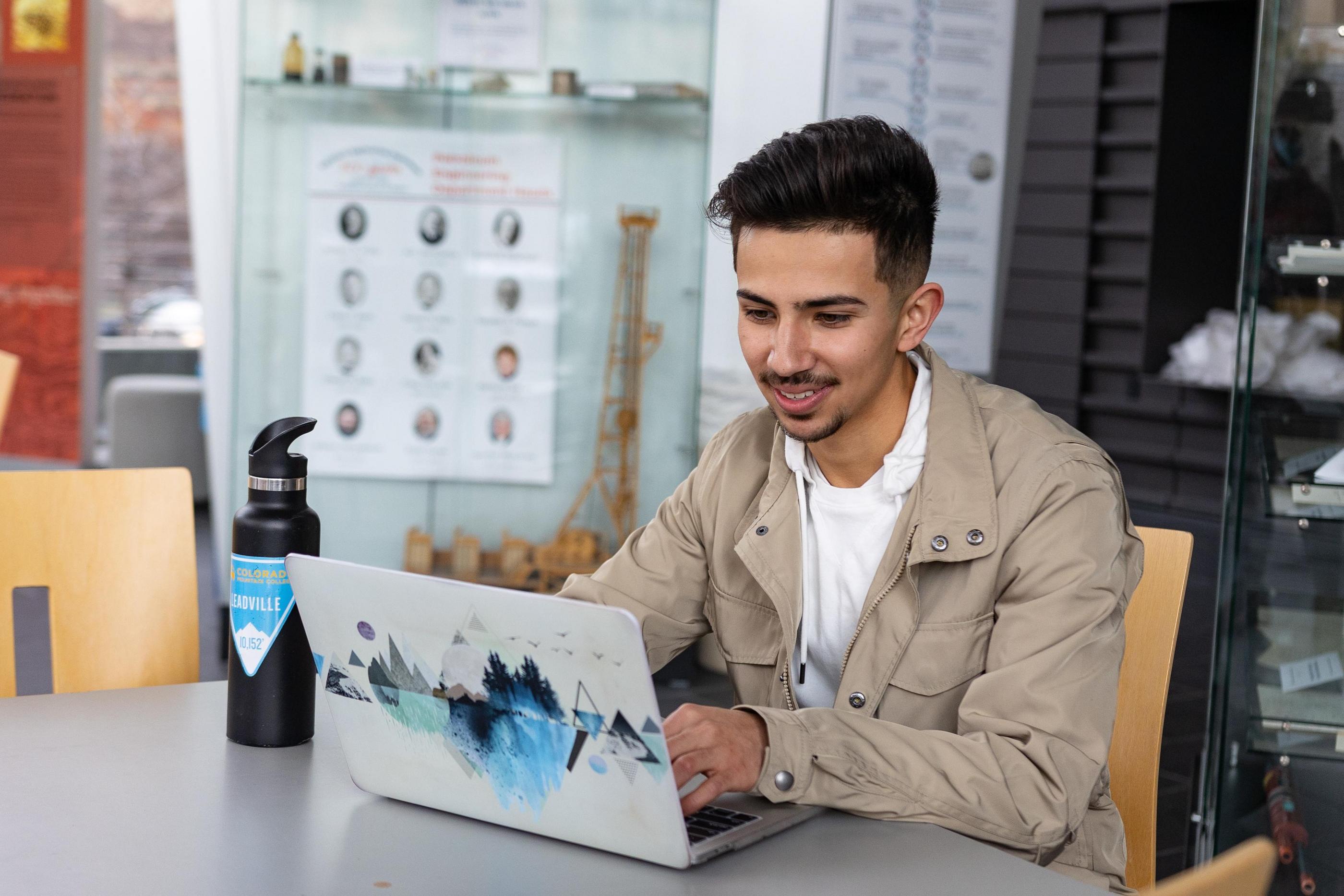 Male student in beige jacket working on a laptop with a geometric mountain design, seated at a table with a black water bottle nearby.