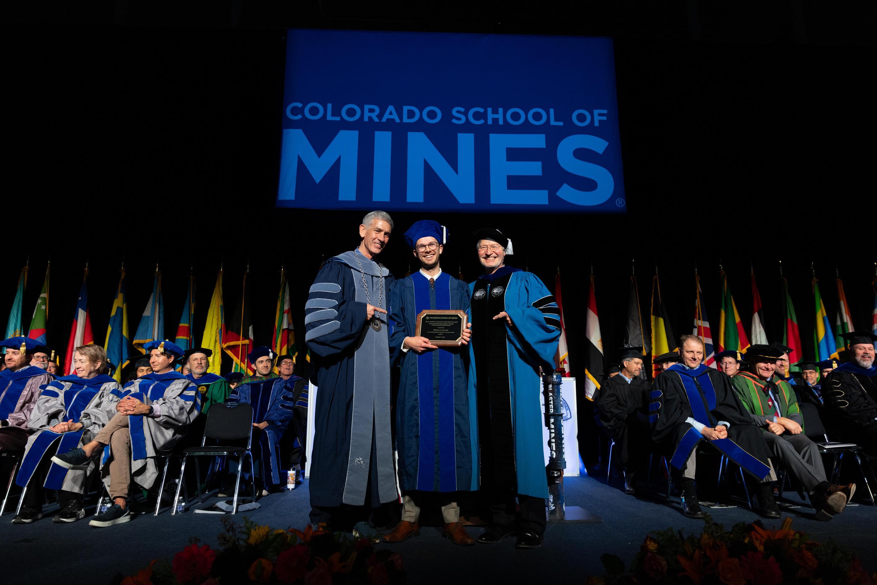 A male PhD graduate stands holding an award between Mines President and Vice President for Research on commencement stage