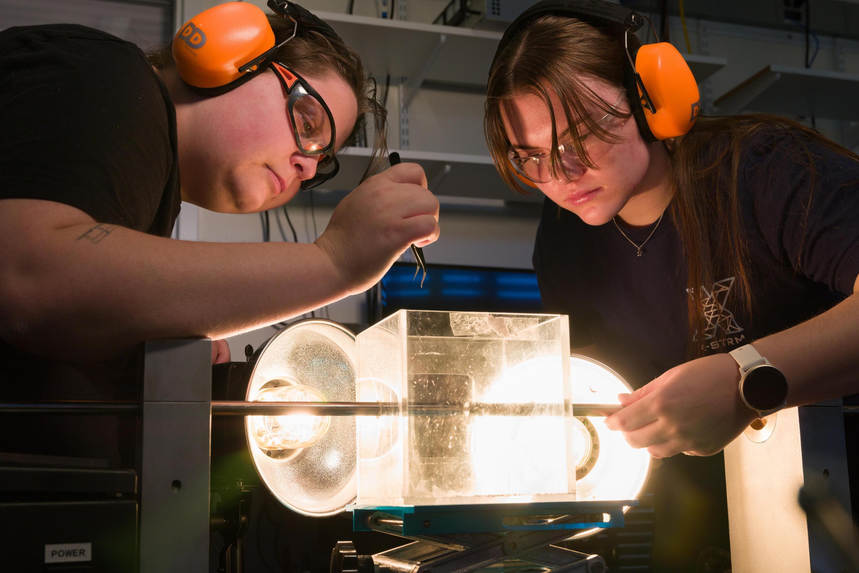 two students in protective eyewear and orange earmuffs lean in around translucent glowing plastic box with metal rod through it