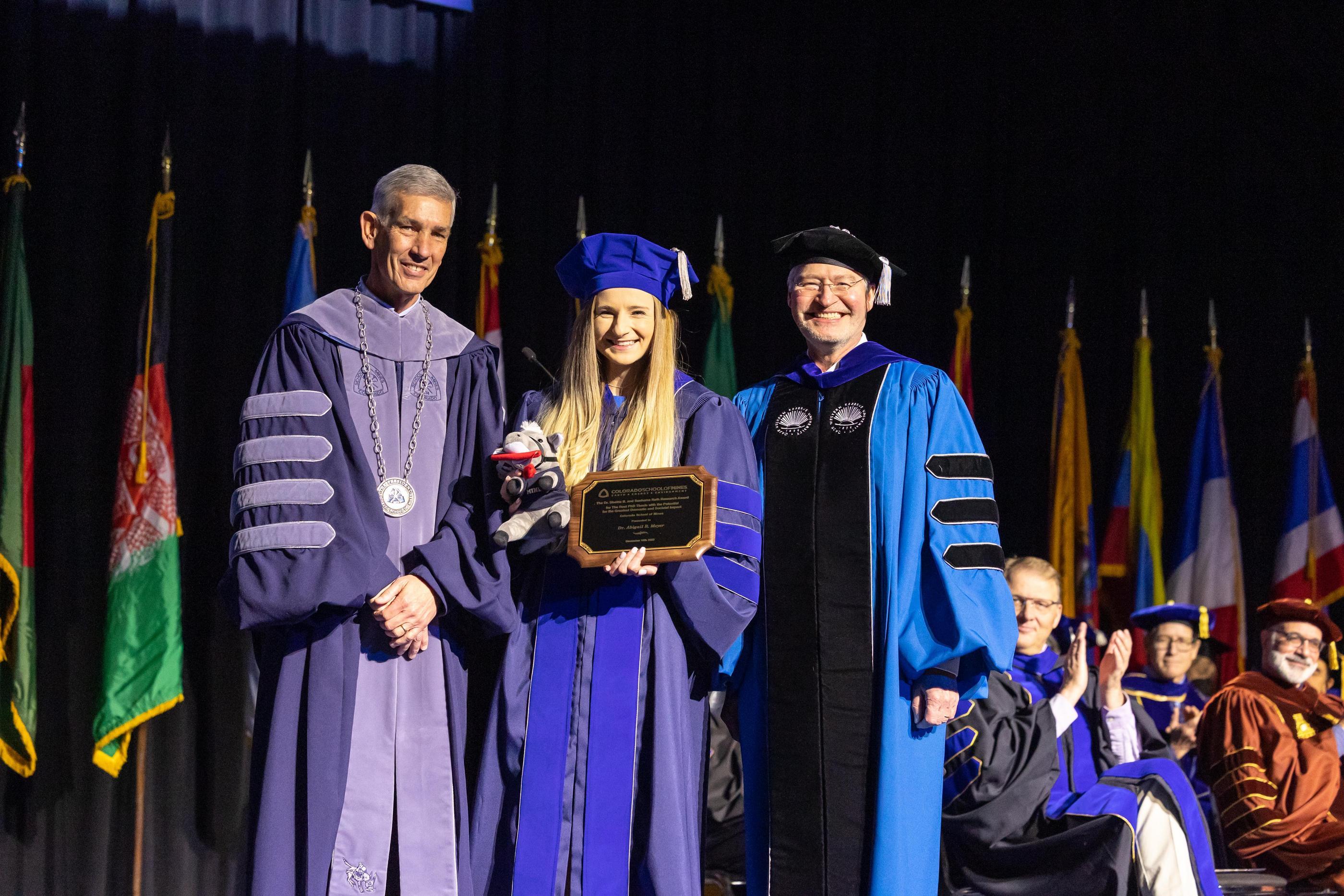 A PhD gradaute holds a plaque between the MInes president and VP of research on Commencement stage