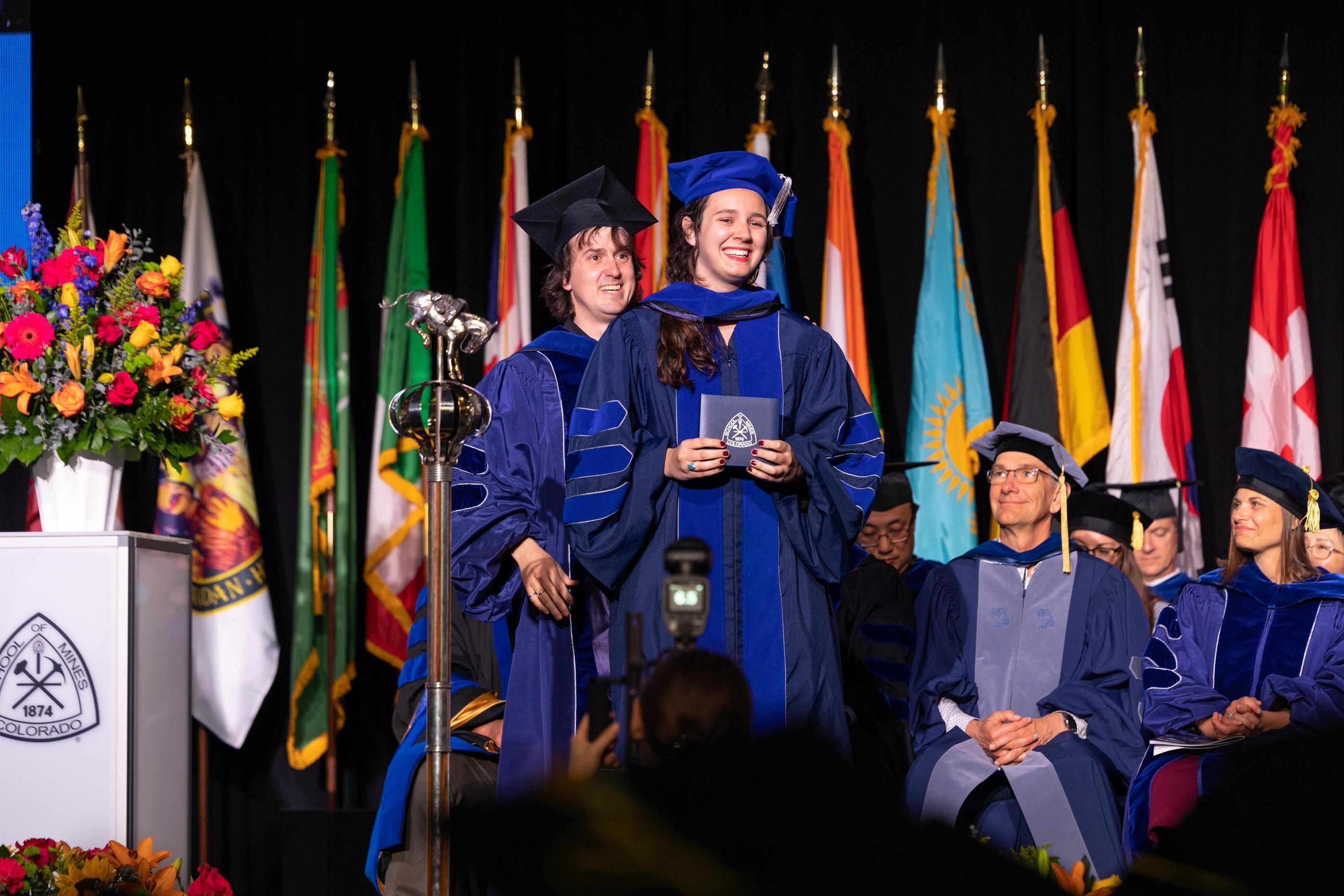 Graduates in blue academic regalia on stage during a commencement ceremony with colorful flags and flowers in the background.