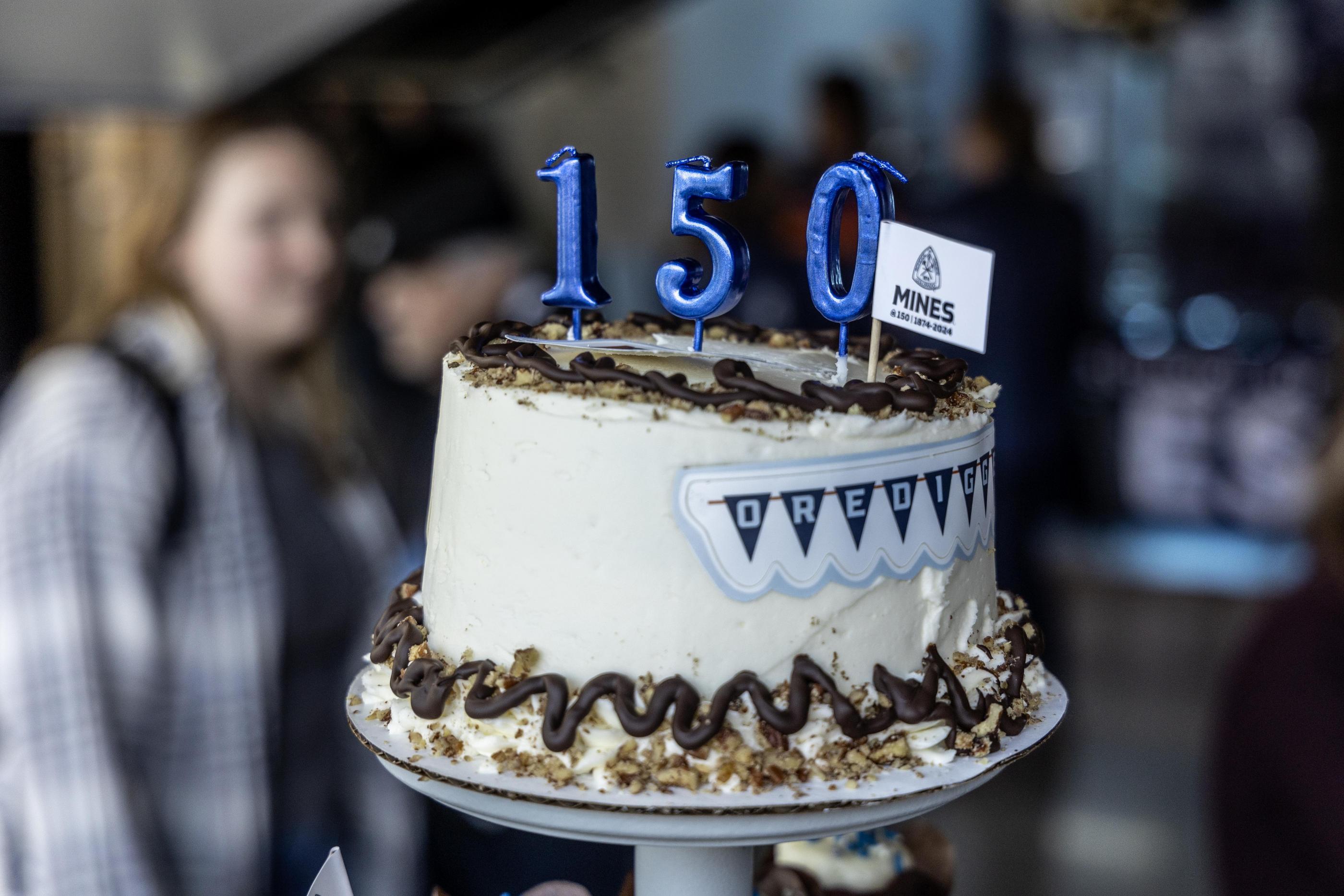 A white birthday cake on a platter with candles in the shape of 150