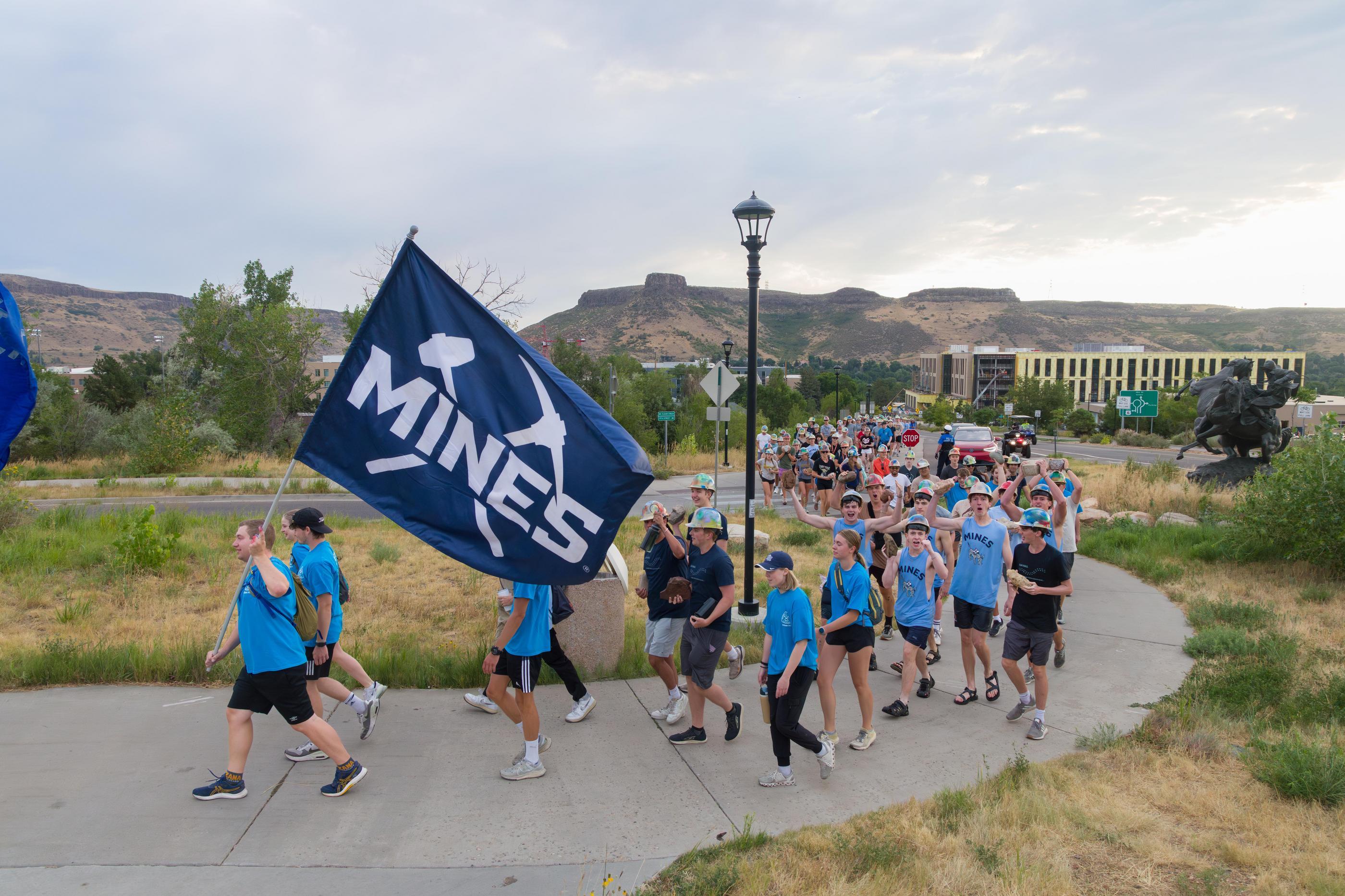students in blue t-shirts walk together on sidewalk with big Mines flag at the front
