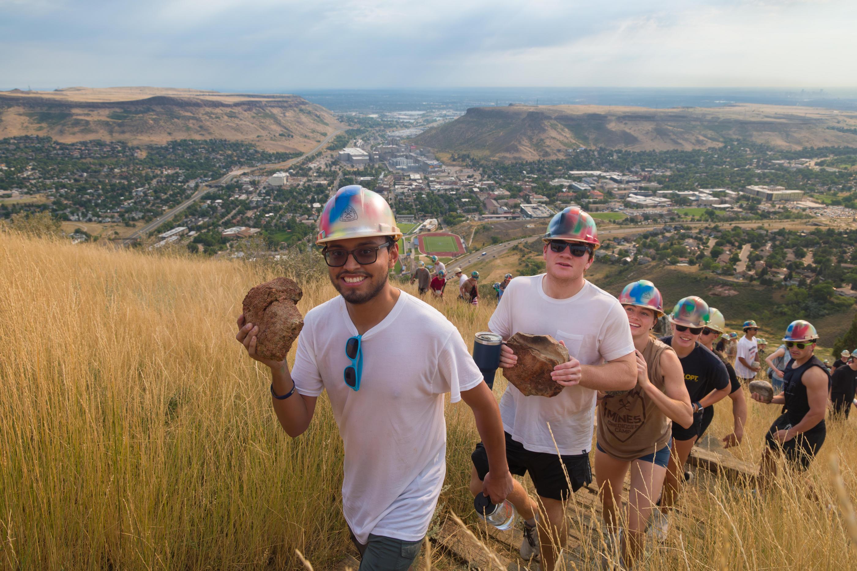 Line of students holding rocks and wearing hard hats climb up a grassy hill with Mines campus visible below
