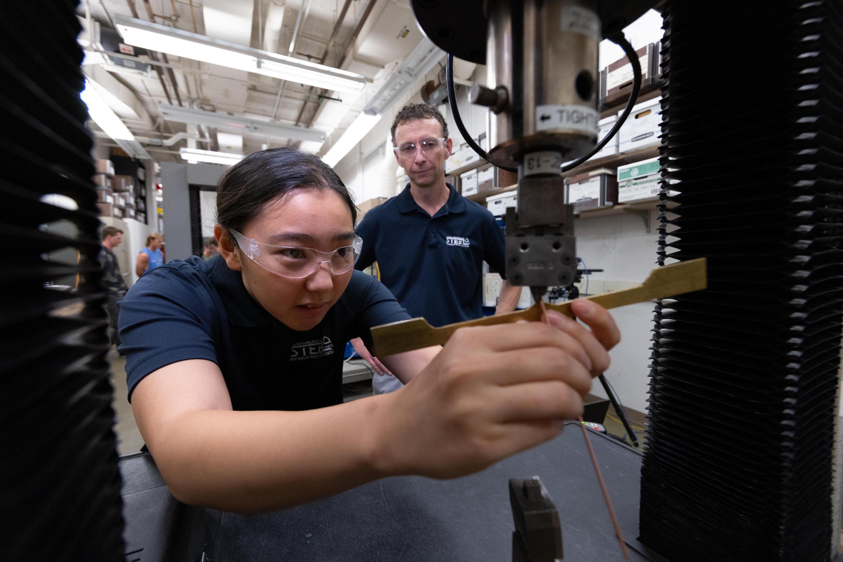 Kip Findley stands behidn a student researcher putting a steel sample into a testing rig