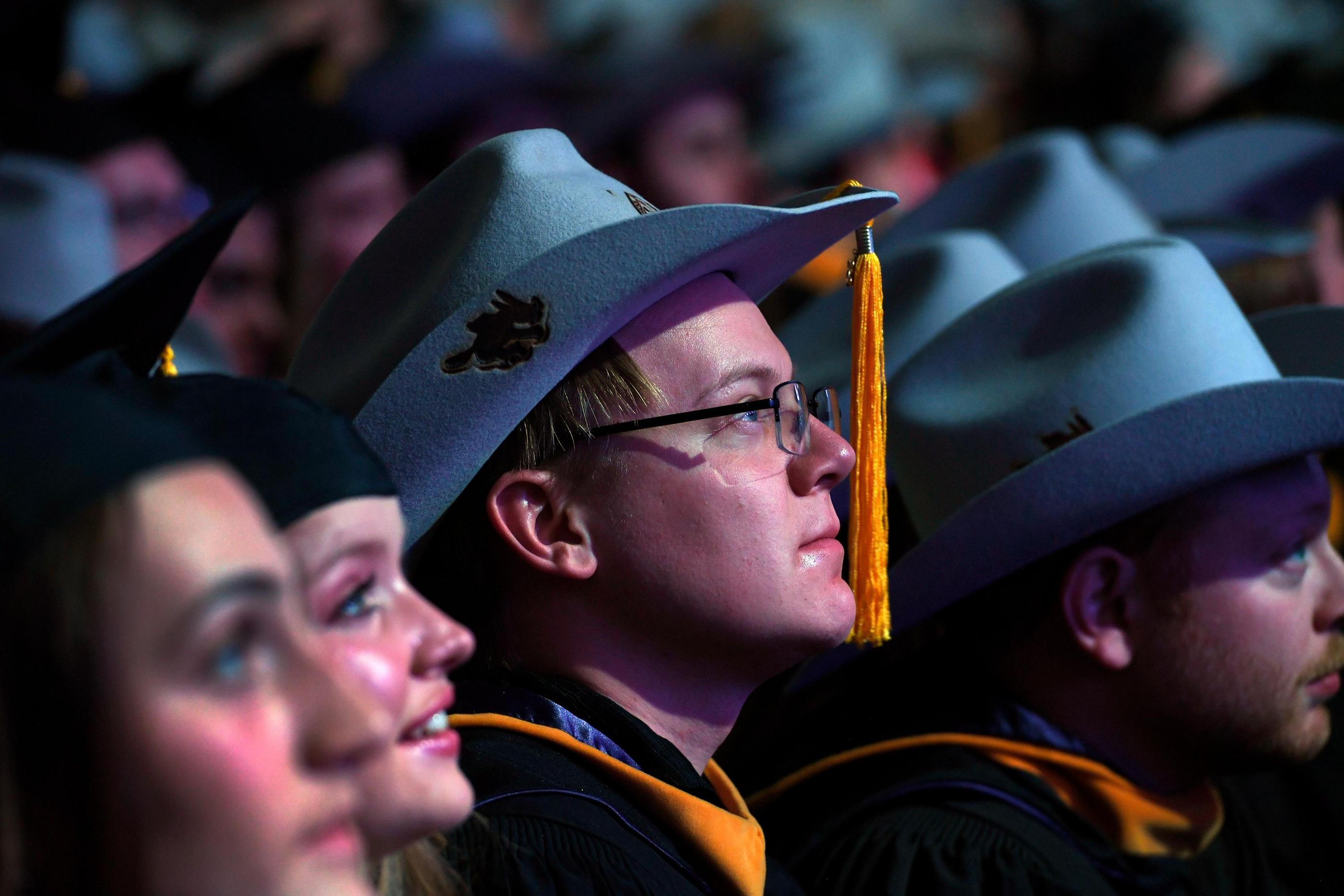Mines graduate student wearing a felt Orediggers cowboy hat with gold tassel listens during commencement, surrounded by fellow graduates in academic regalia.