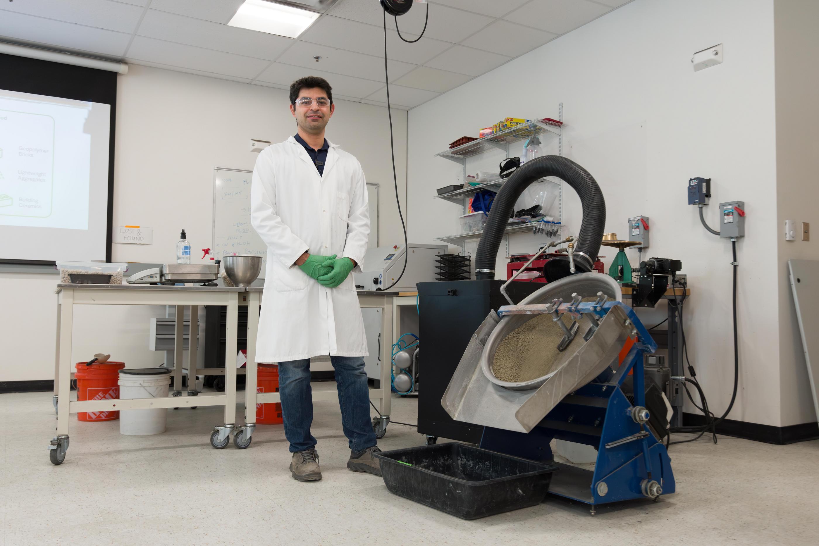 Reza Hedayat wears a lab coat, safety glasses and green gloves next to a large machine in the mine tailings lab.