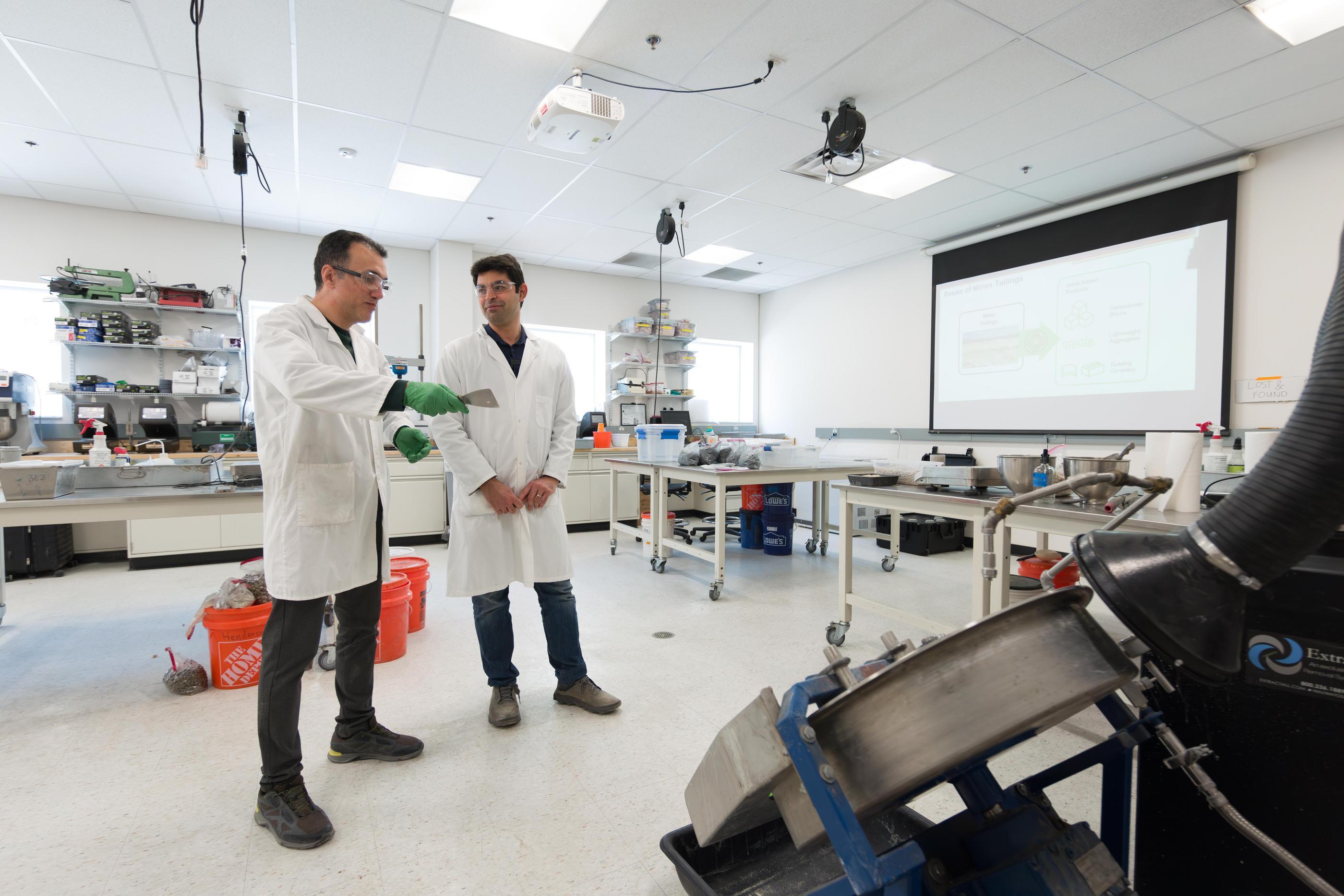Reza Hedayat, right, and Farid Shabani stand in a Colorado School of Mines research lab as Shabani holds a mine tailings sample, with lab equipment and a presentation screen in the background.