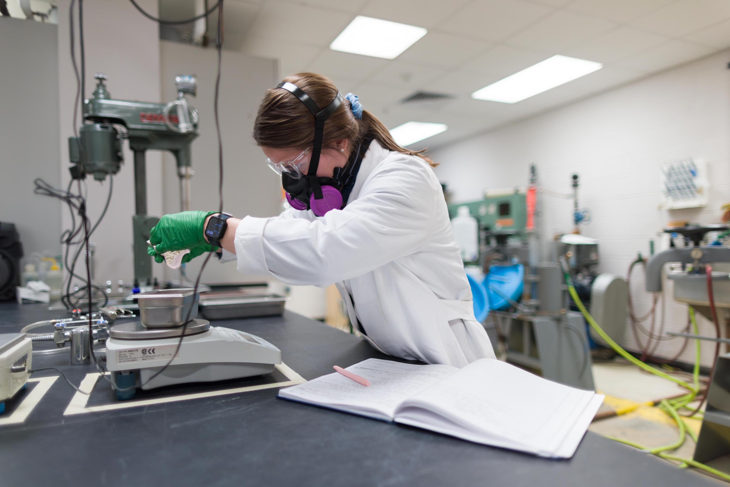 A Mines PhD student in safety glasses and a respirator weighs out a sample in a lab.