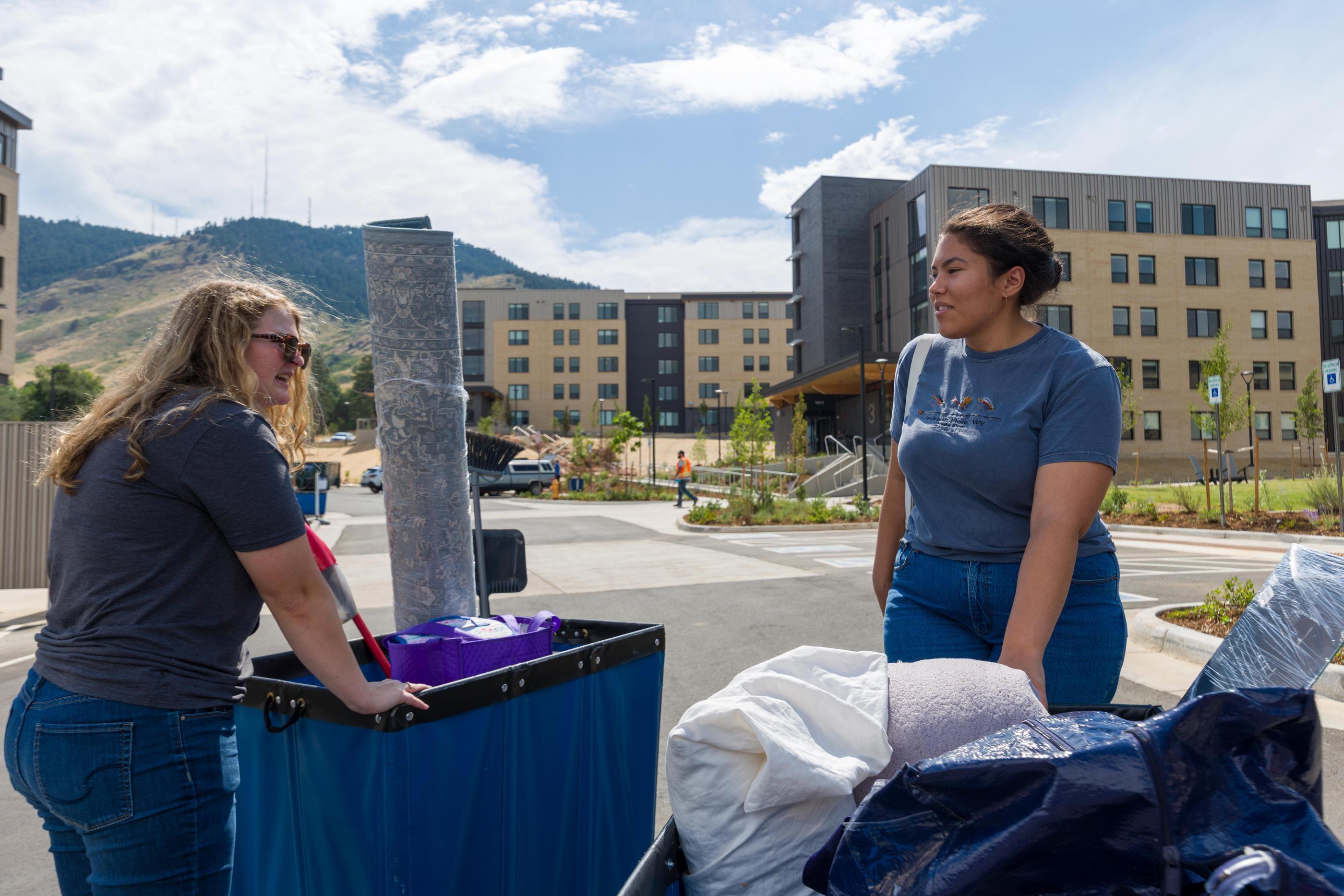 Two female students push moving carts with Mines Park buildings and blue sky in background