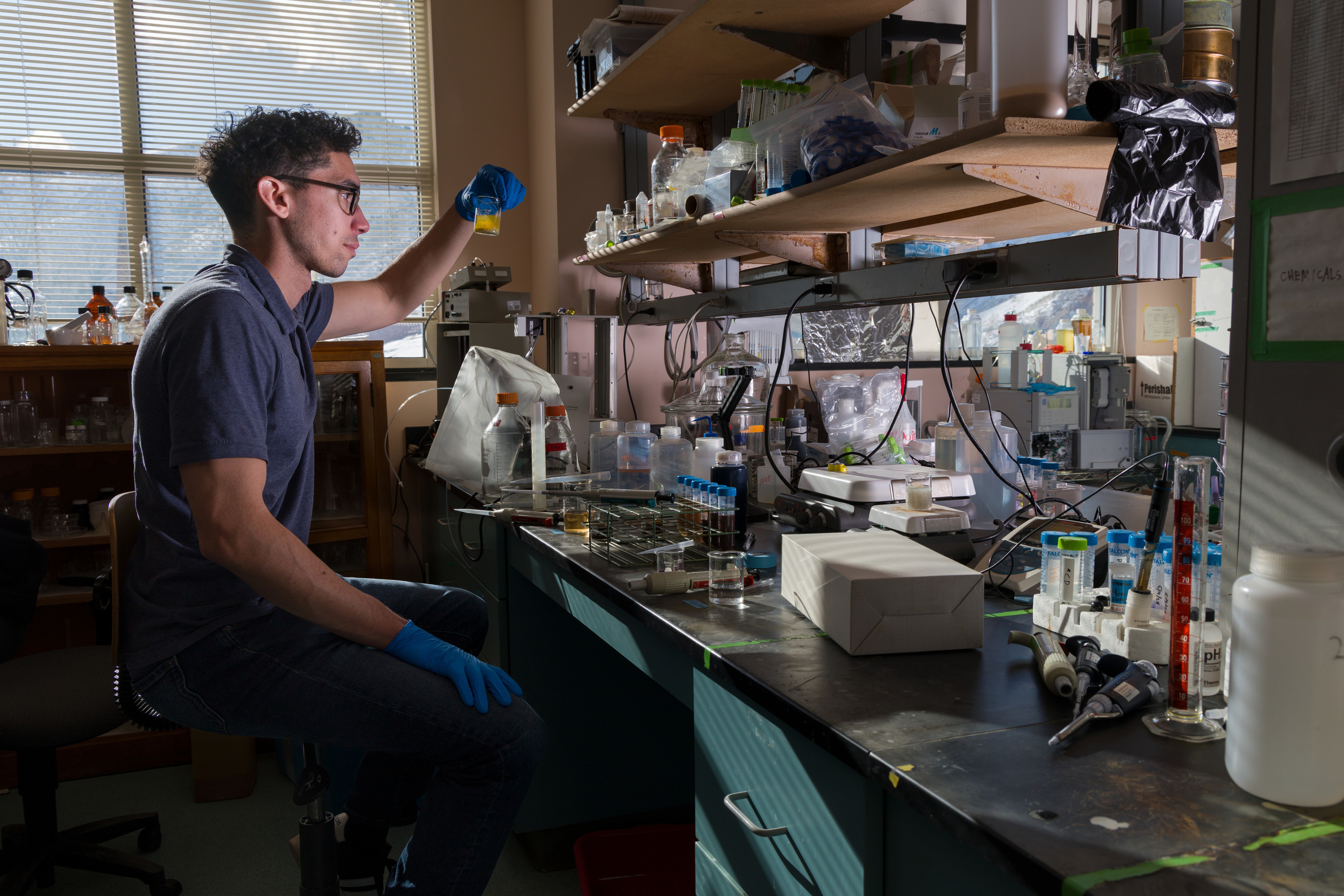 Male researcher sits at lab bench looking at vial held up in his hand