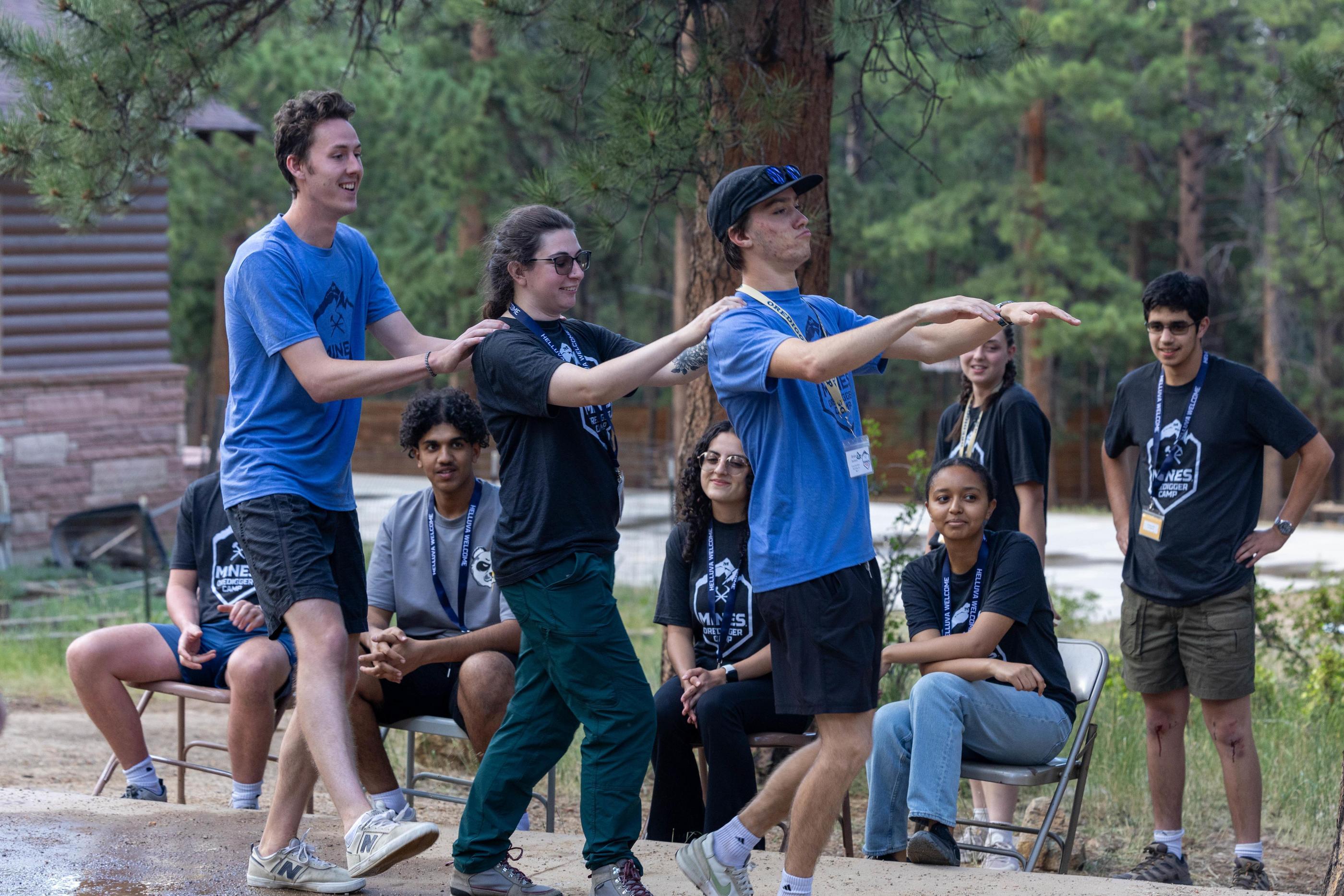 Three students perform a skit in the woods with other students looking on