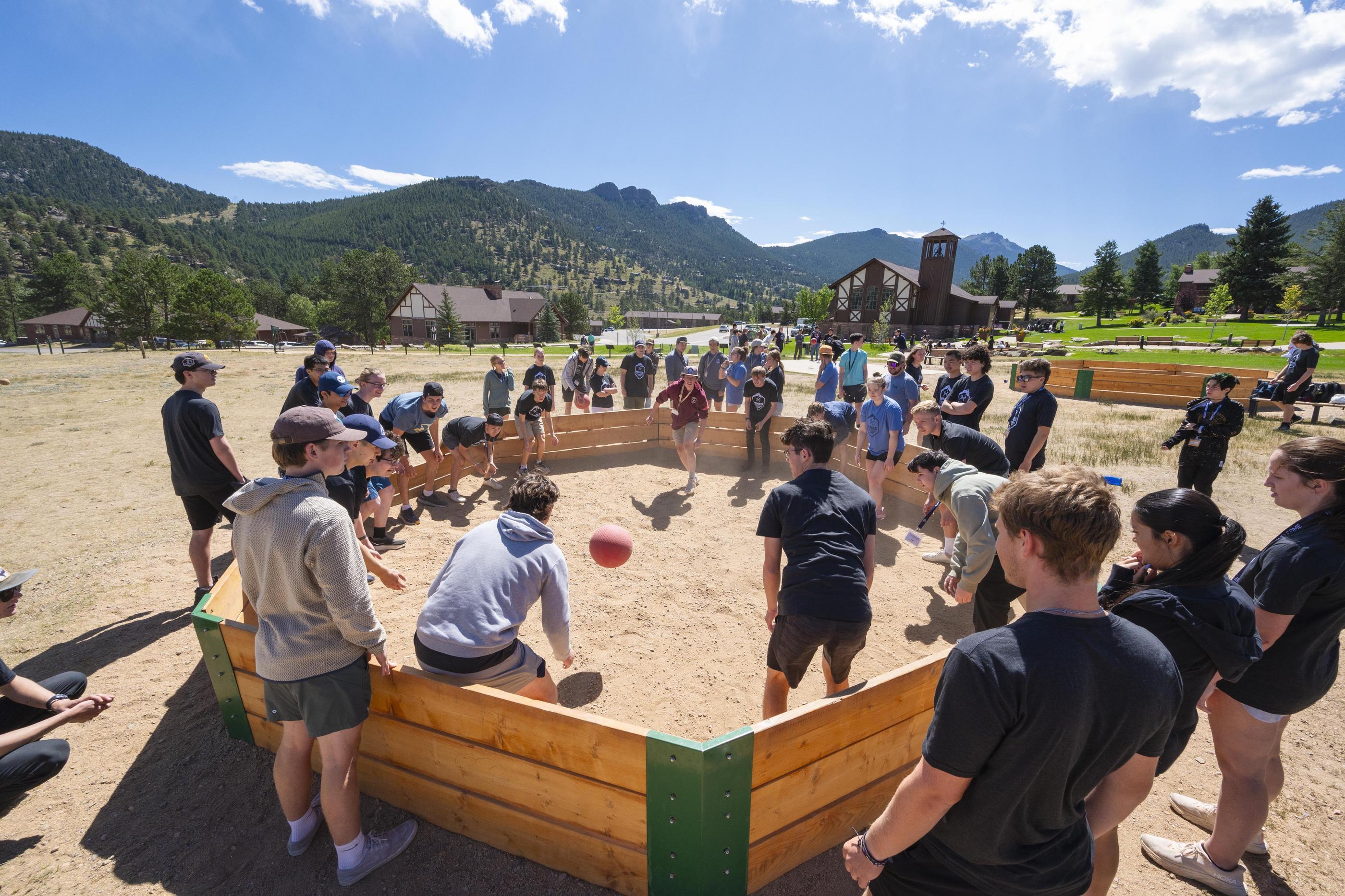 A large group of first-year students plays Gaga Ball in a wooden pit during Oredigger Camp, with mountains and camp buildings in the background at the YMCA of the Rockies.