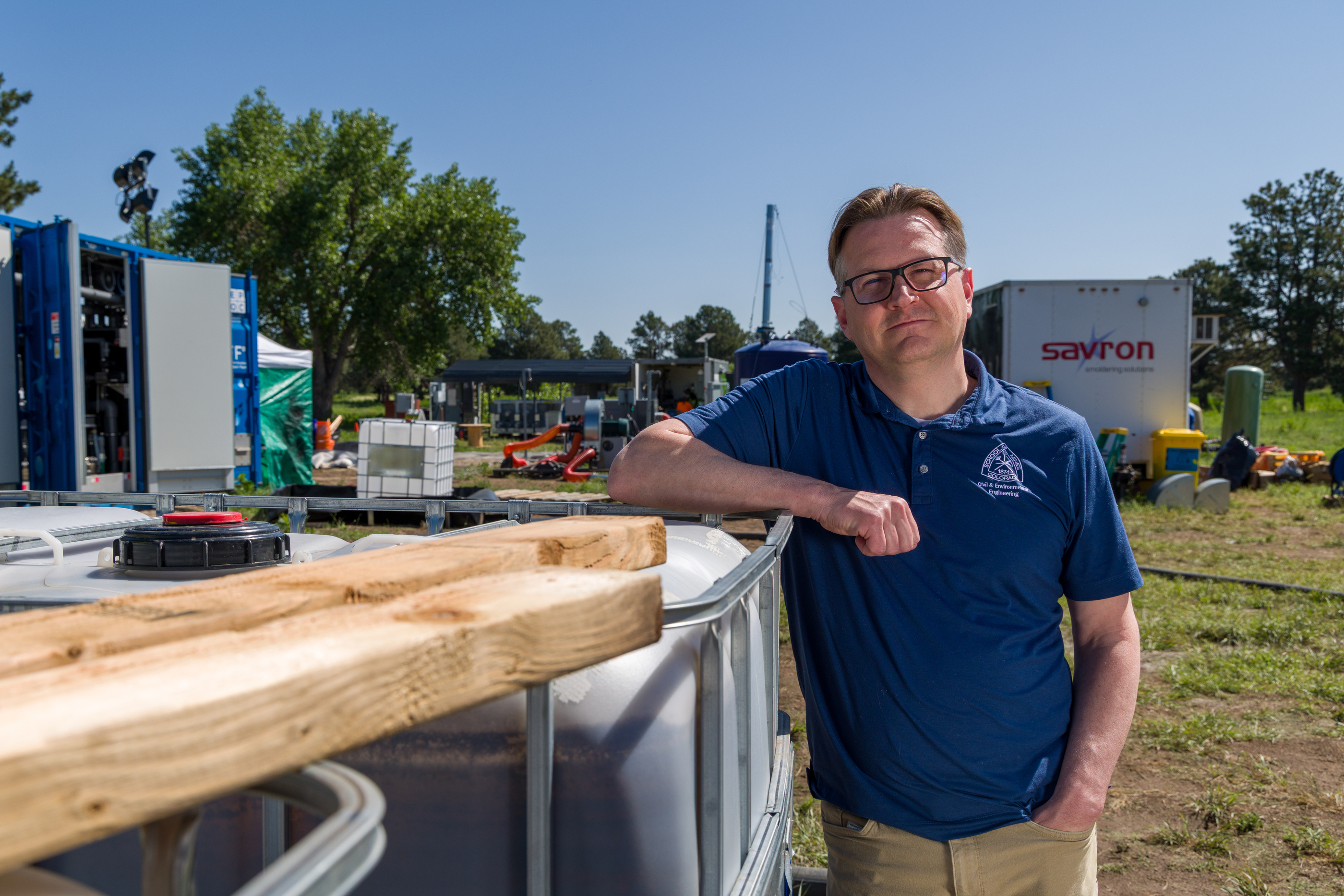 Chris Higgins, a Colorado School of Mines Civil and Environmental Engineering researcher, stands at a PFAS water treatment testing site, leaning on equipment tanks with mobile treatment units in the background.