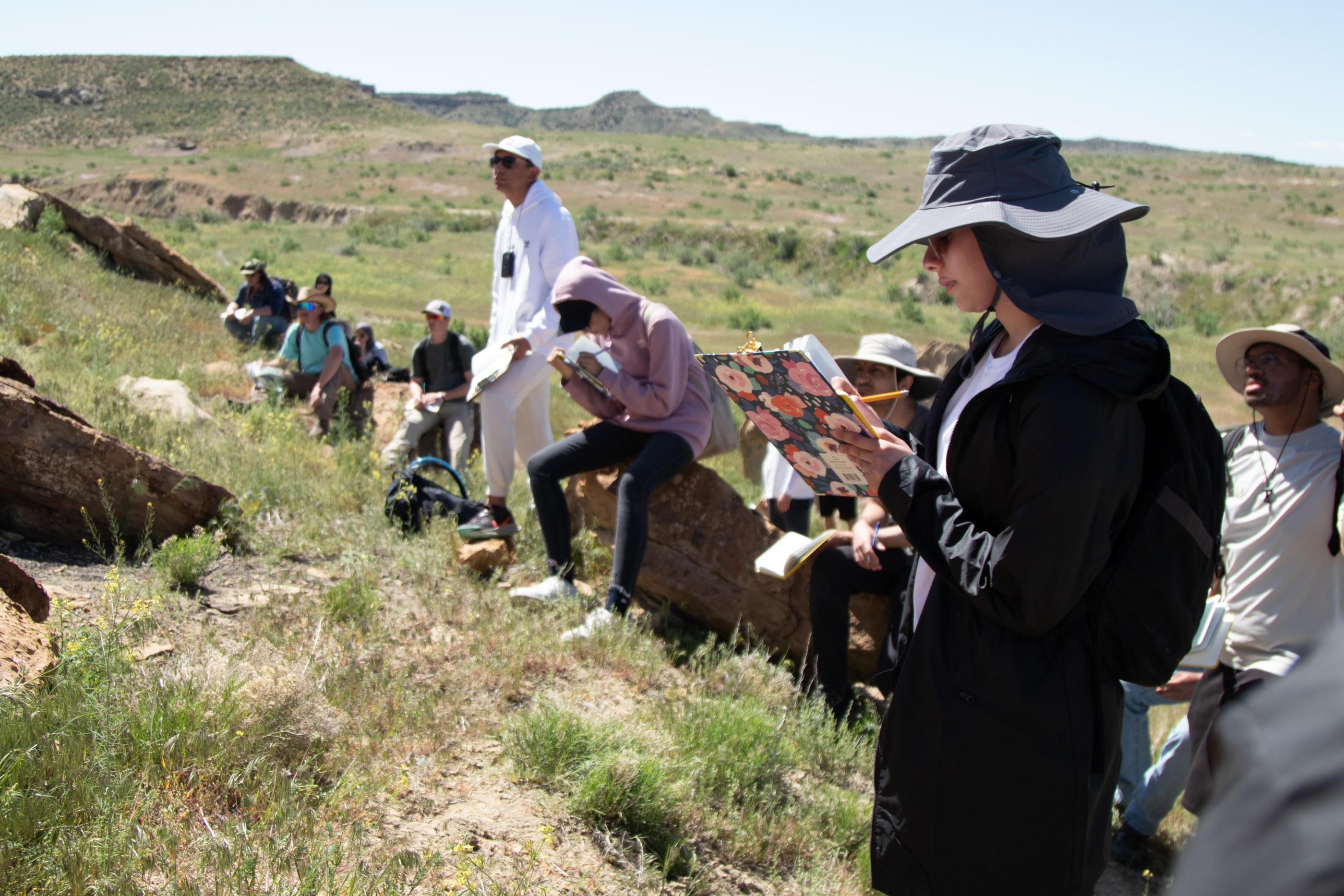Students sit and stand in a field taking notes