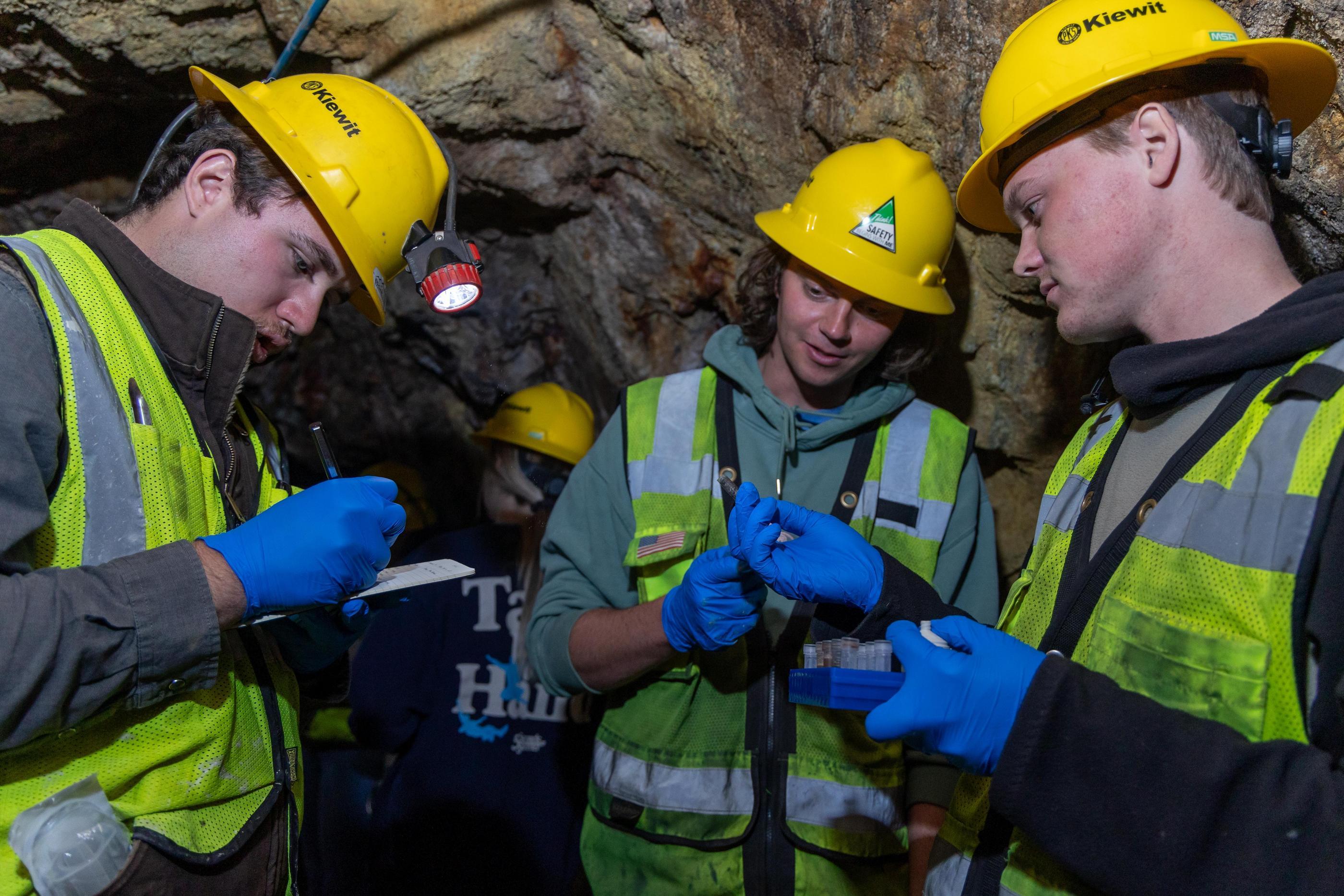 Three students in hard hats and head lamps take notes on sample underground
