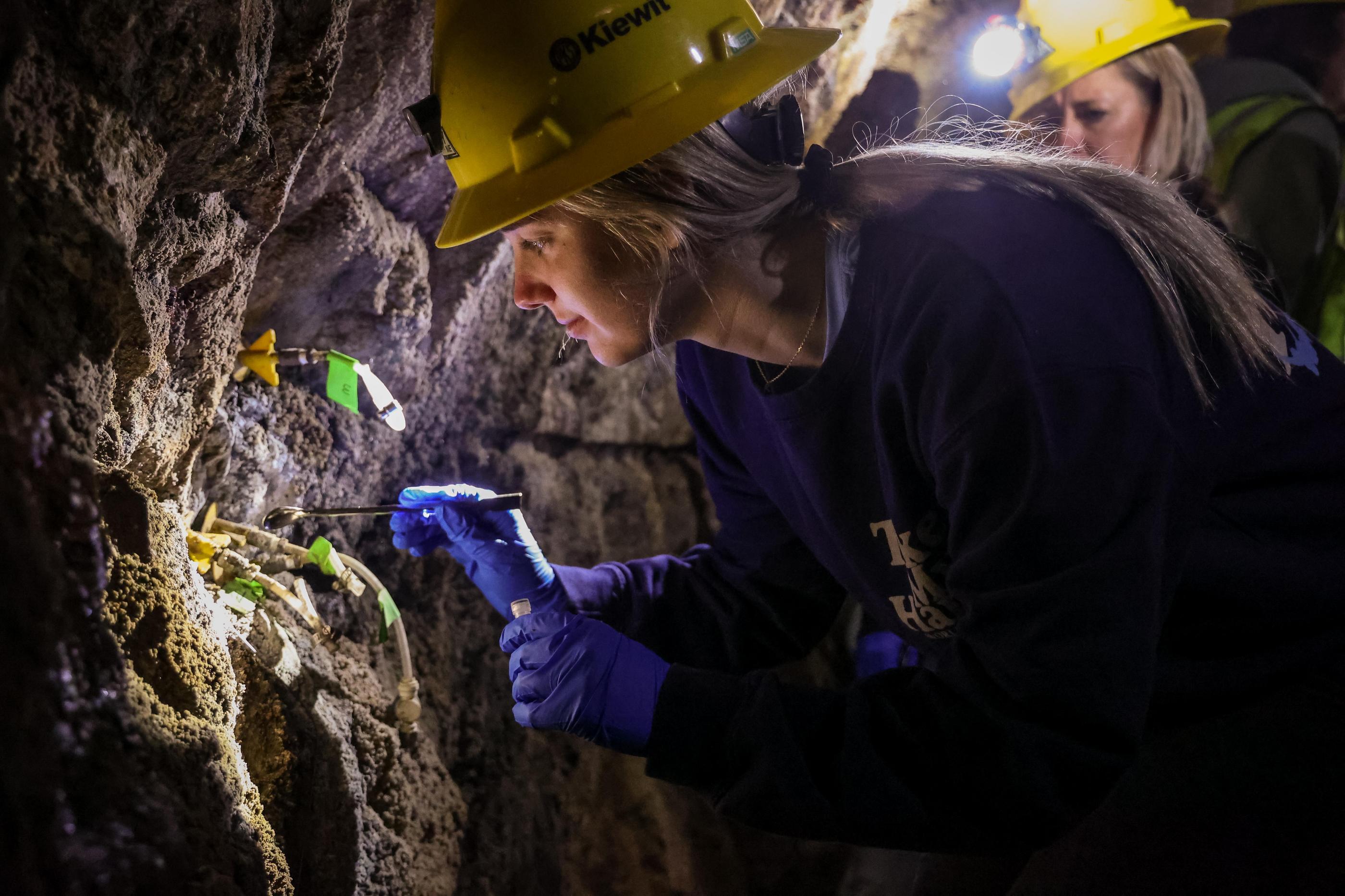 A student in yellow hard hat and head lamp takes a sample from a rock wall inside the Edgar Mine