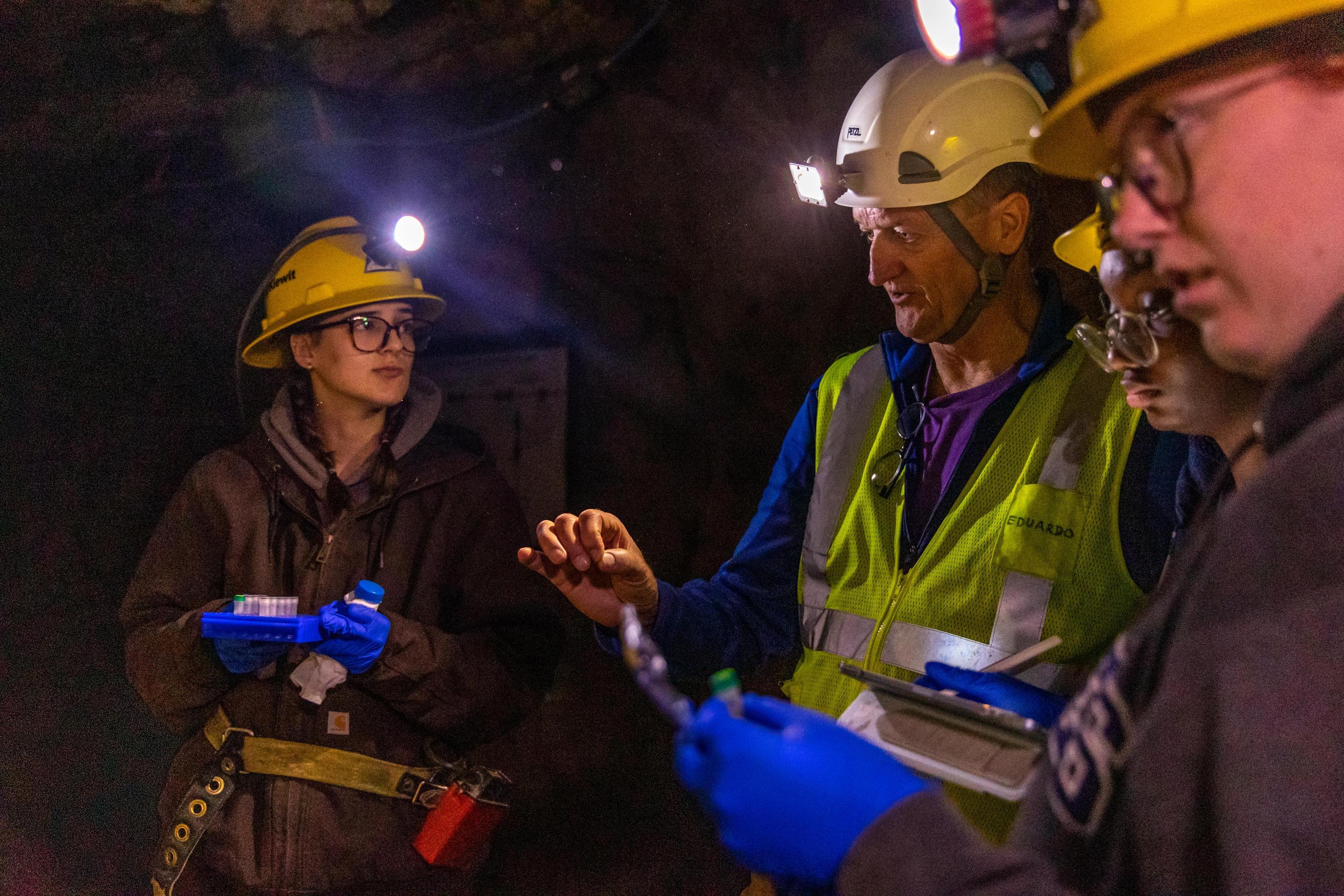 A professor and two students in yellow hard hats and head lamps examine a sample underground