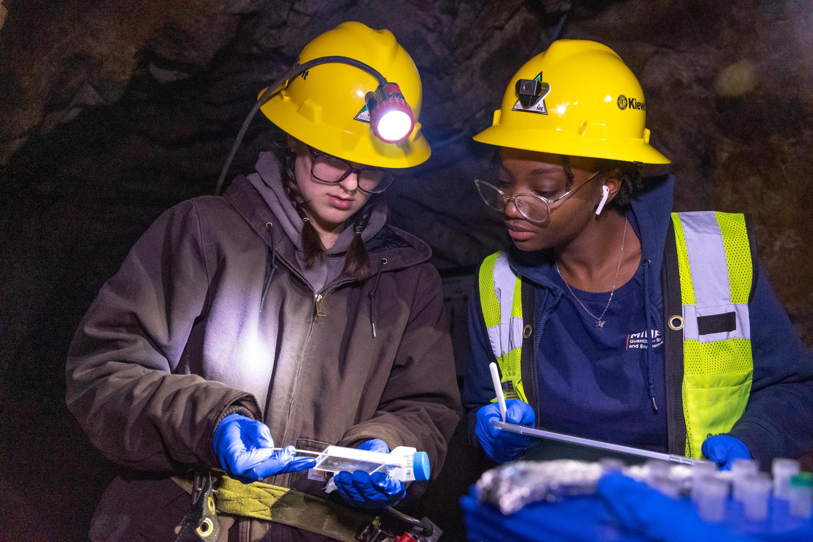 Two students in yellow hardhats and head lamps look at sample results in the mine