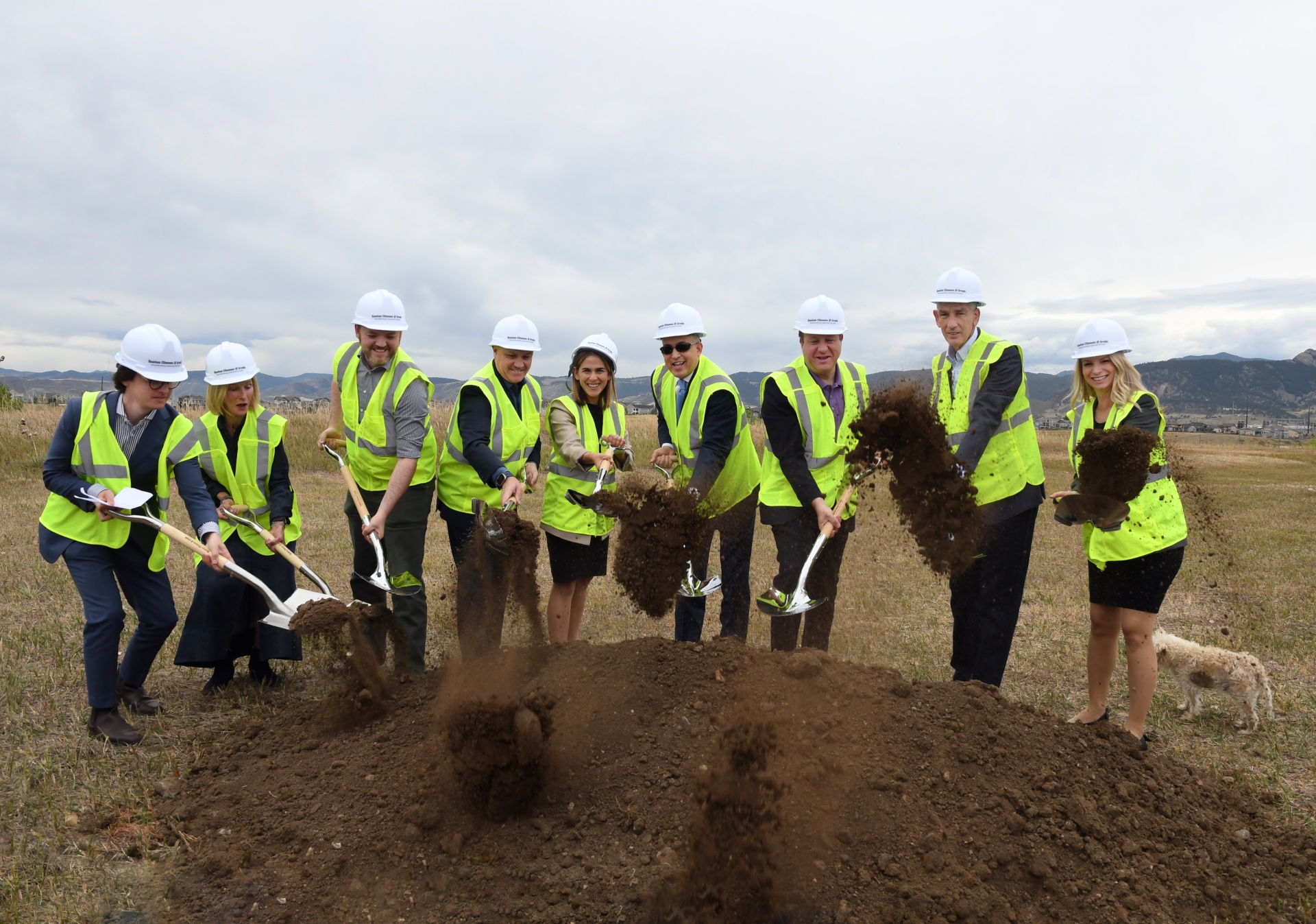 A line of dignitaries in yellow safety vests and white hard hats scoop ceremonial dirt into the air