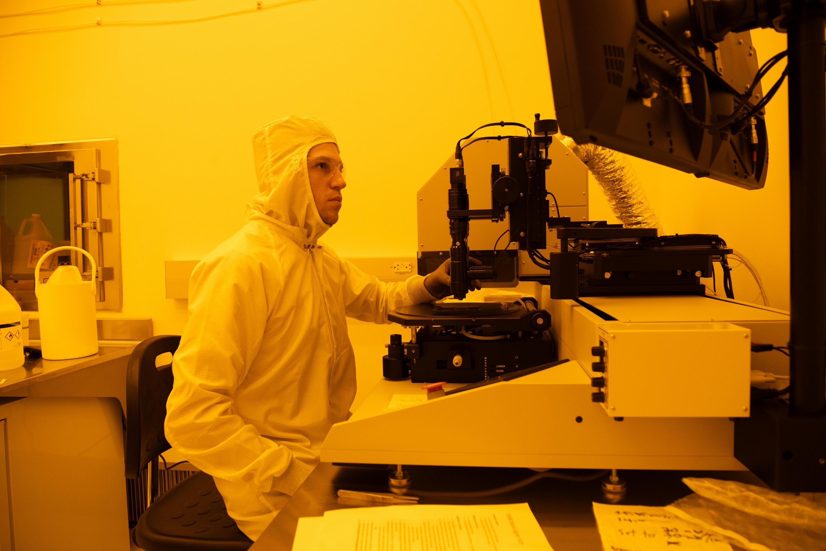 A student wearing a white bunny suit sits at a piece of lab equipment in a clean room