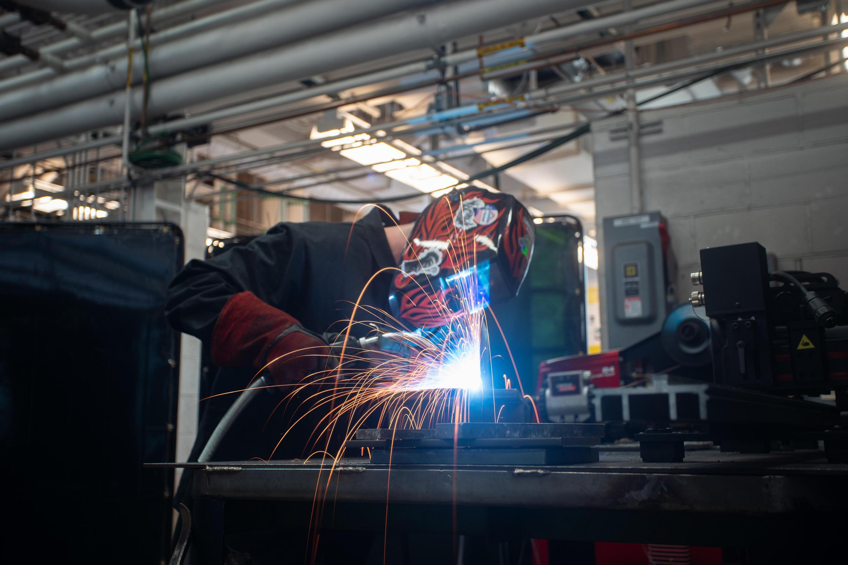 A student wearing a red helment welds a piece of metal, causing a spray of bright sparks.