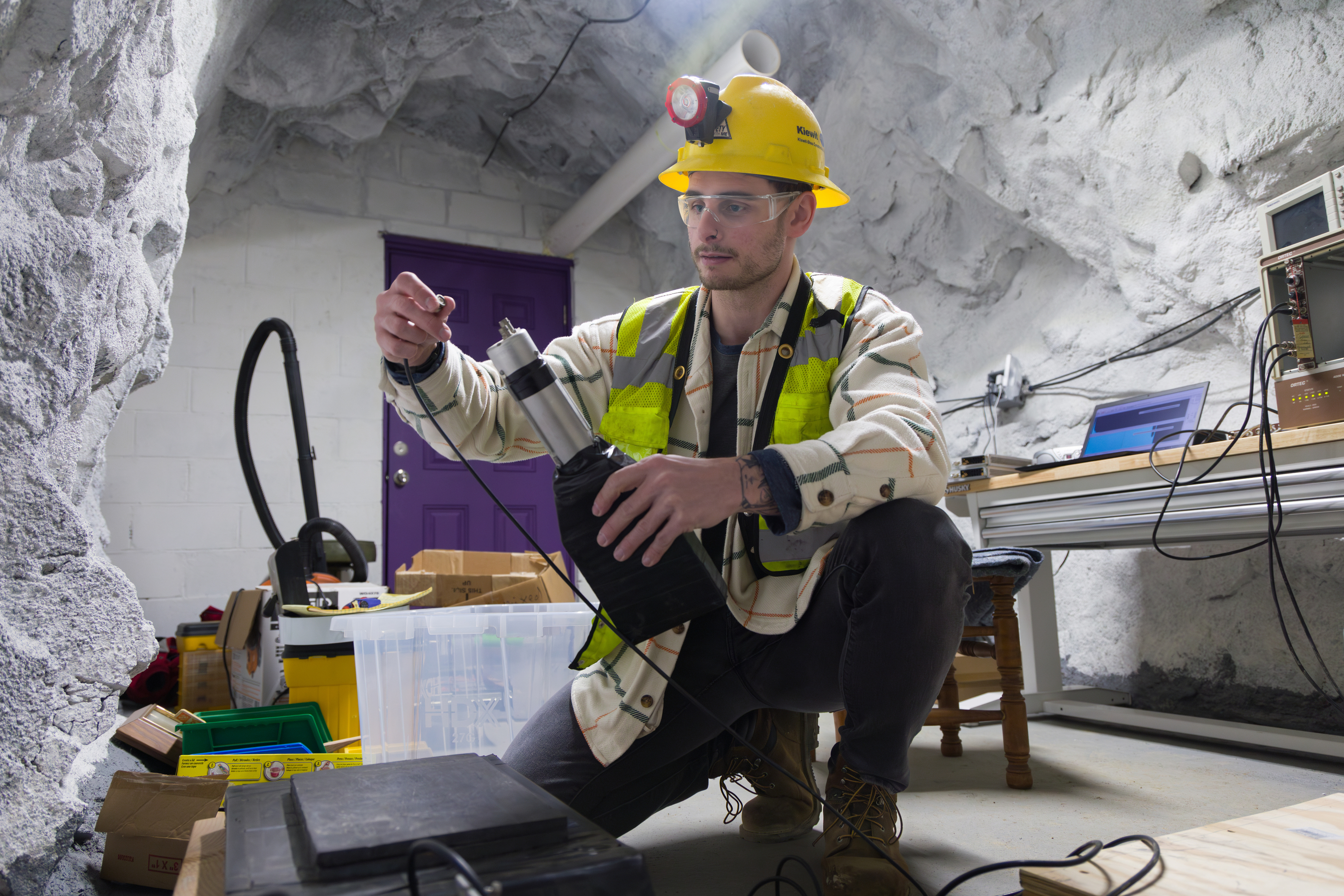 A student in a hard hat and safety glasses assembles a device in an underground lab space.
