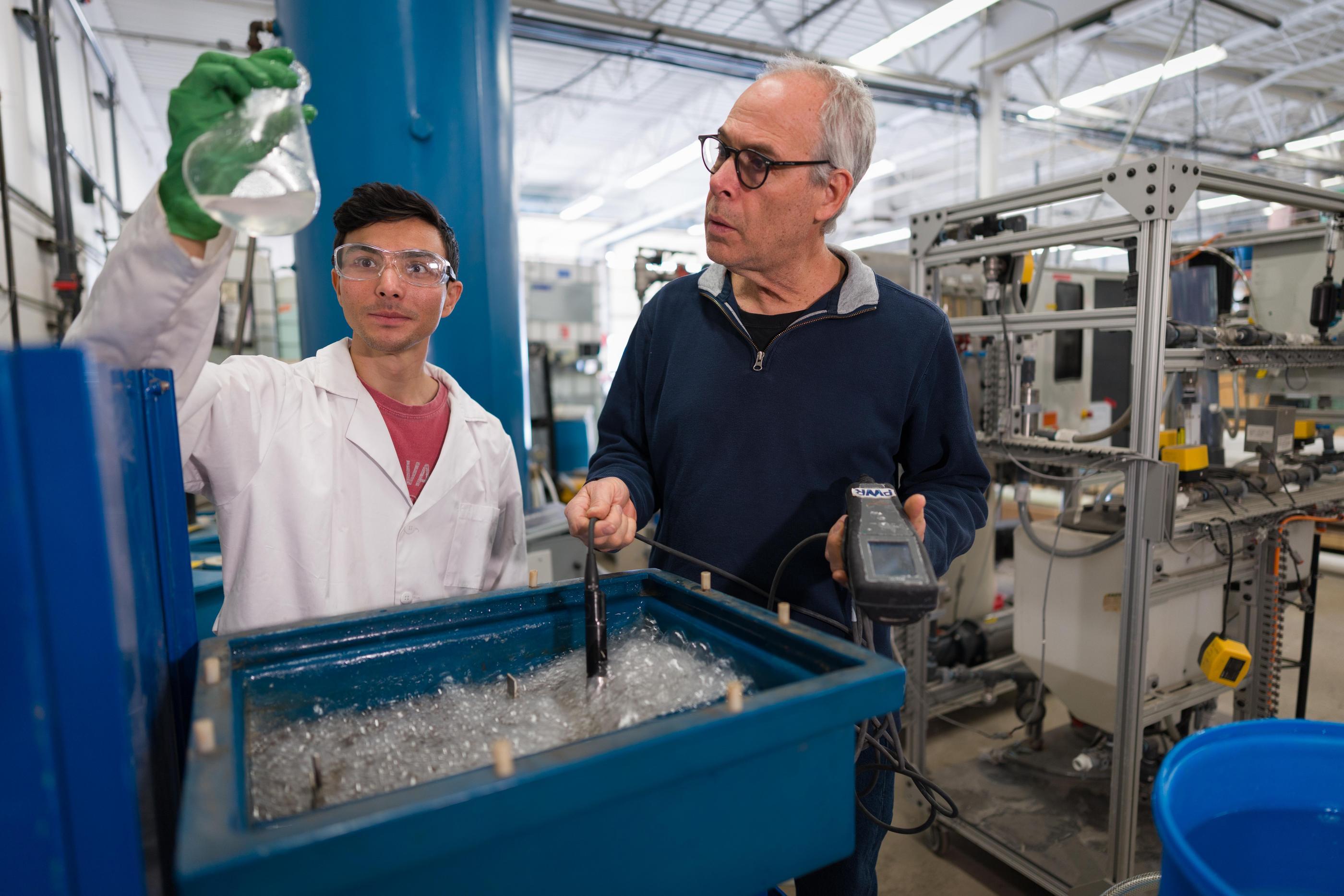 A graduate student wearing safety glasses holds up a flask of clear liquid while an older professor dips a probe into a tub of aerated, bubbling water in a testing lab.