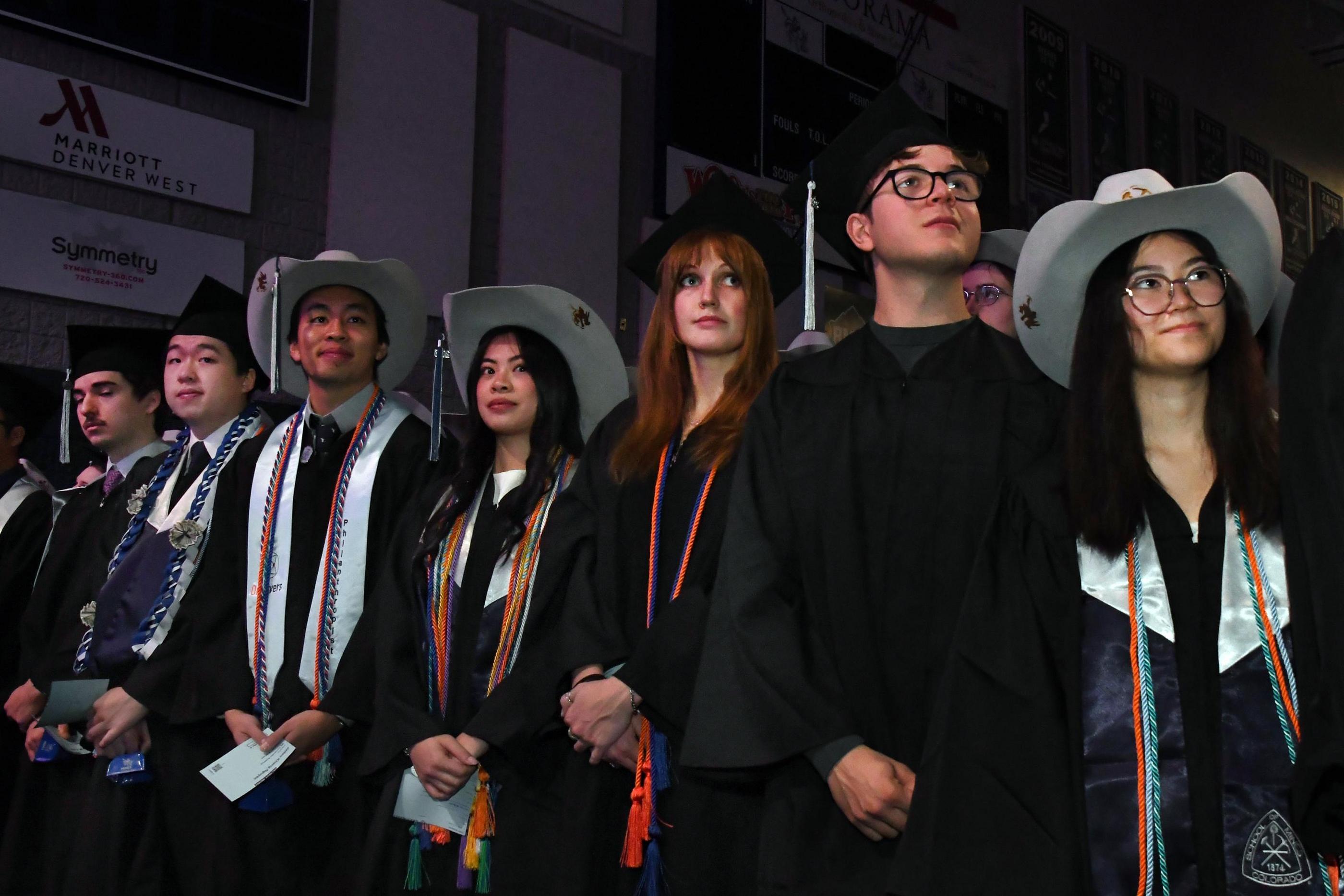 Mines undergraduate students in caps, gowns and honor cords, some wearing Orediggers cowboy hats, stand together awaiting the start of commencement.