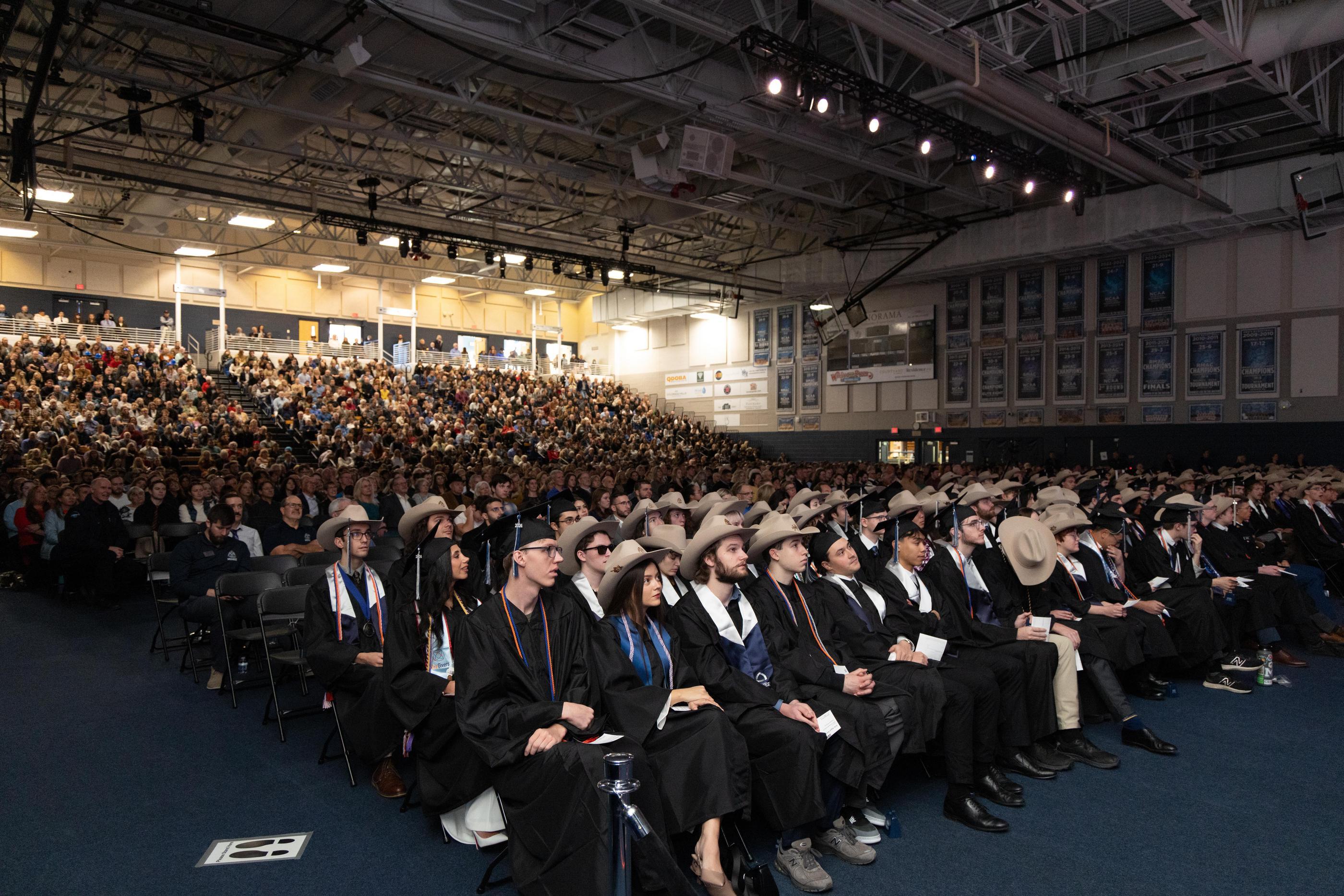 Colorado School of Mines undergraduates in academic regalia and traditional cowboy hats sit in the foreground during commencement, with a packed crowd of family and guests filling the arena behind them.