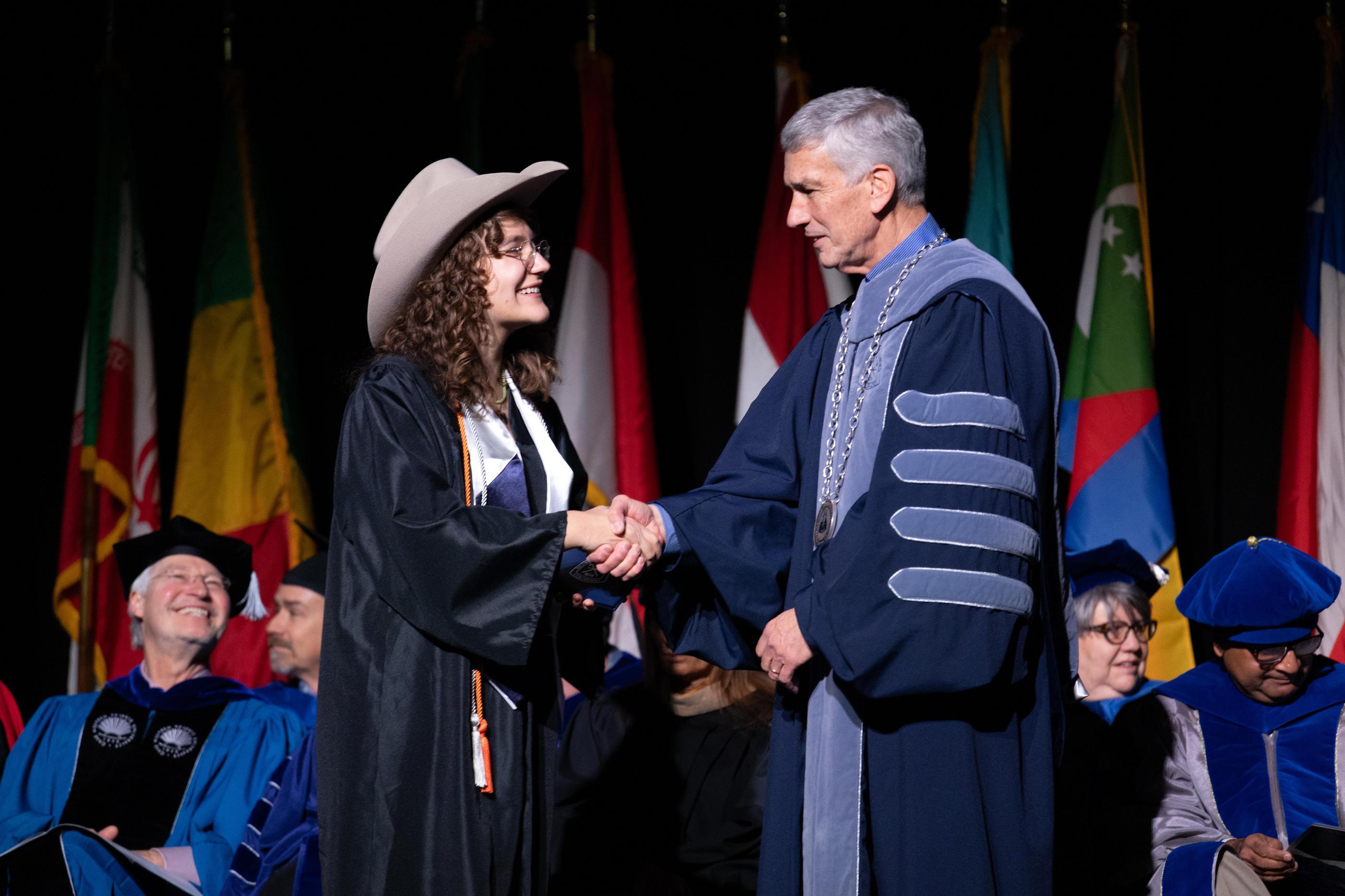 An undergraduate student in a Mines hat shakes President Johnson's hand on commencement stage