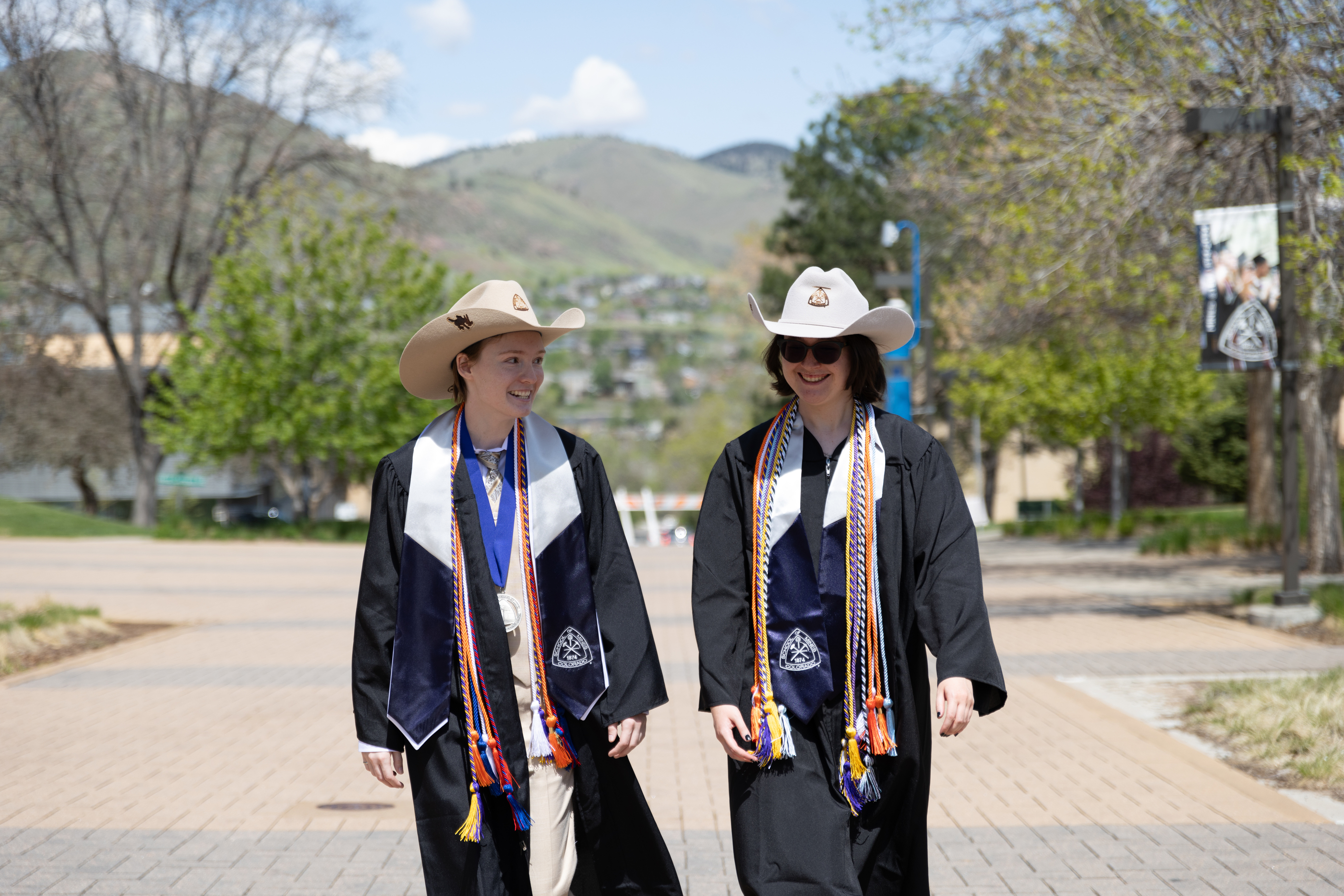 Two undergraduate students in commencement regalia and Mines Hats walk down pedestrian plaza together