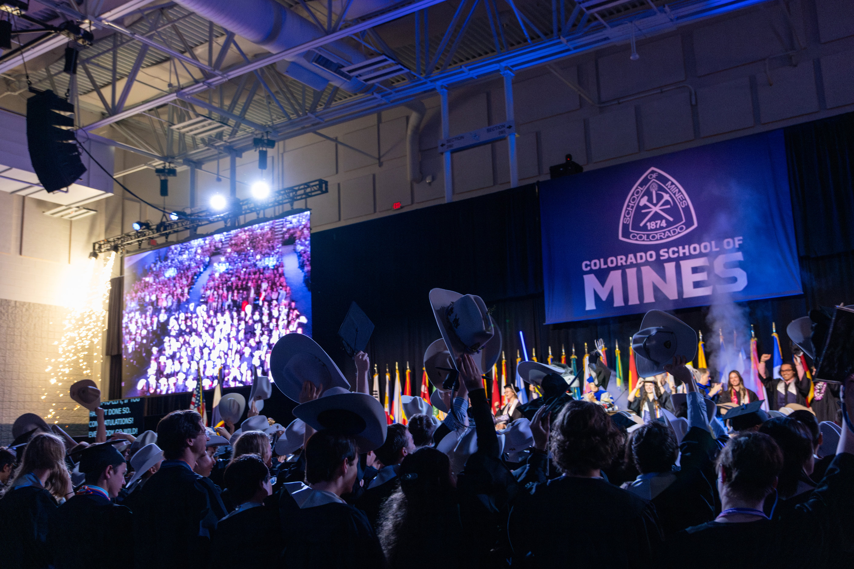 Graduates raise their cowboy hats in celebration during the Colorado School of Mines undergraduate commencement ceremony, with the Mines logo displayed on stage.